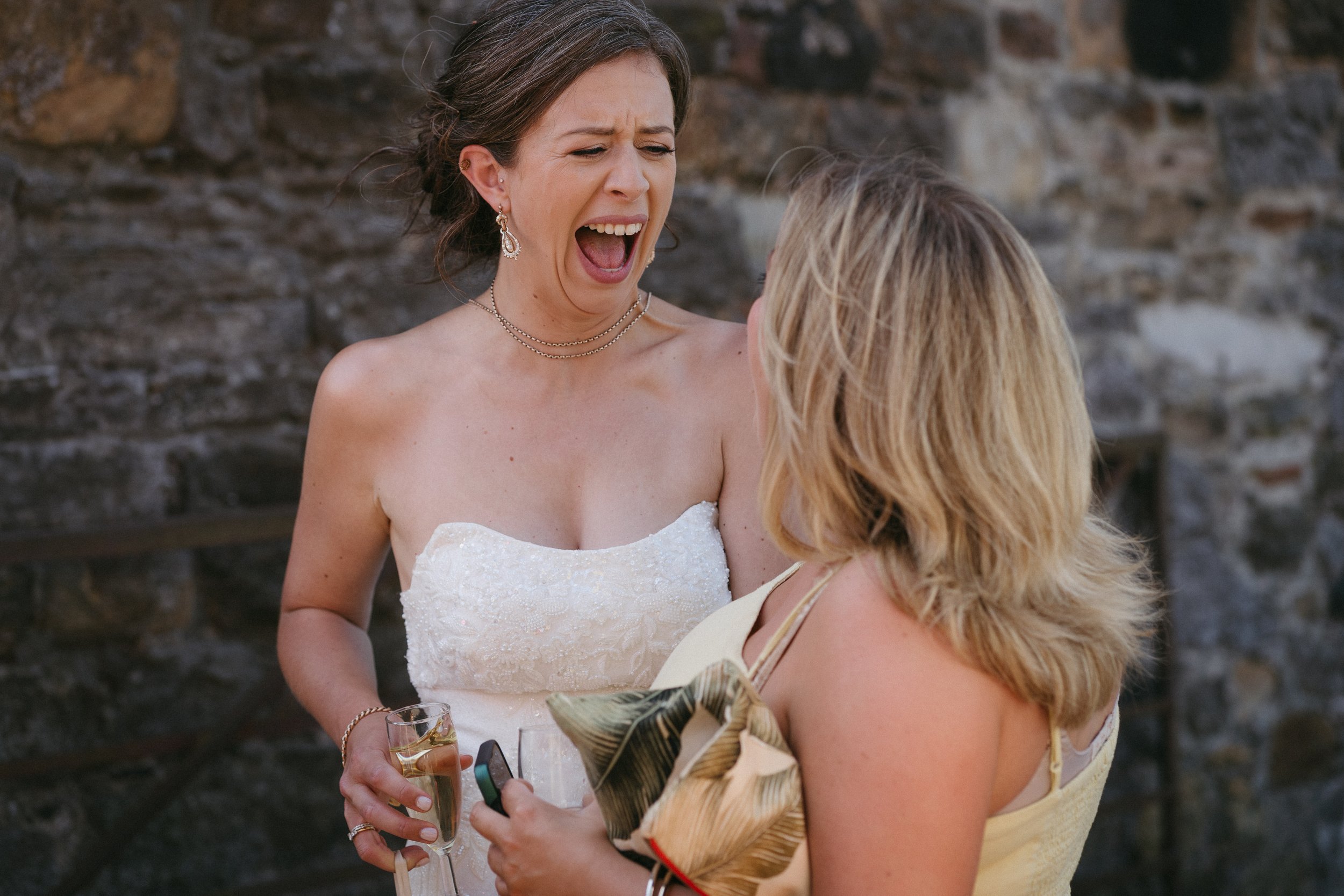 A woman in a white strapless wedding dress is laughing and talking to another woman with blonde hair, wearing a yellow dress. The woman in the wedding dress is holding a glass of champagne. - captured by an Edinburgh wedding photographer