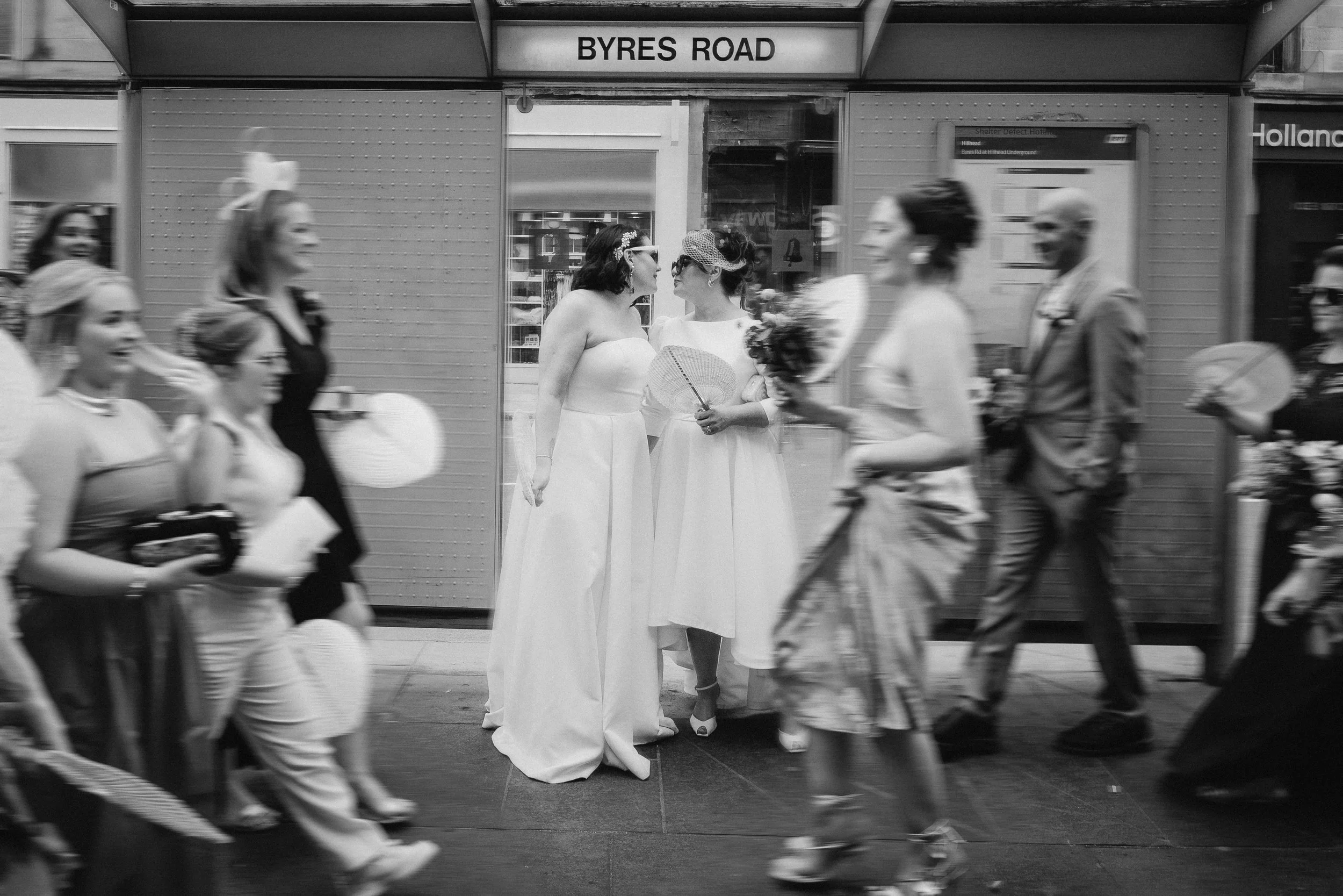 Black and white photo of two women in wedding dresses standing close together among pedestrians on a city sidewalk. The scene is in front of a storefront with a sign that reads "BYRES ROAD." Other people walk past, some holding flowers or umbrellas. 