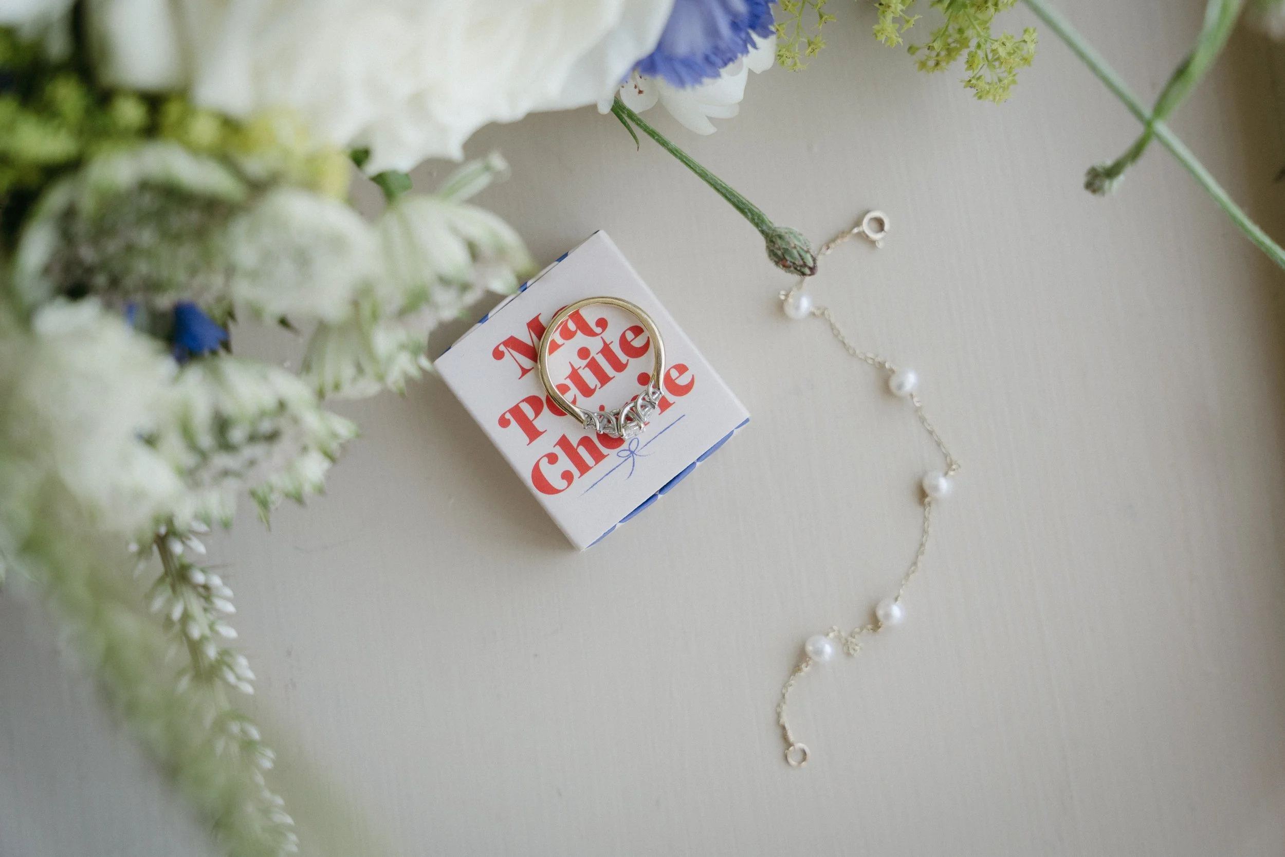 A small white box with red text that reads 'Matte Petite Chrissie' surrounded by white and blue flowers, a silver ring with small gemstones, and a pearl necklace on a light surface. - captured by an Edinburgh wedding photographer