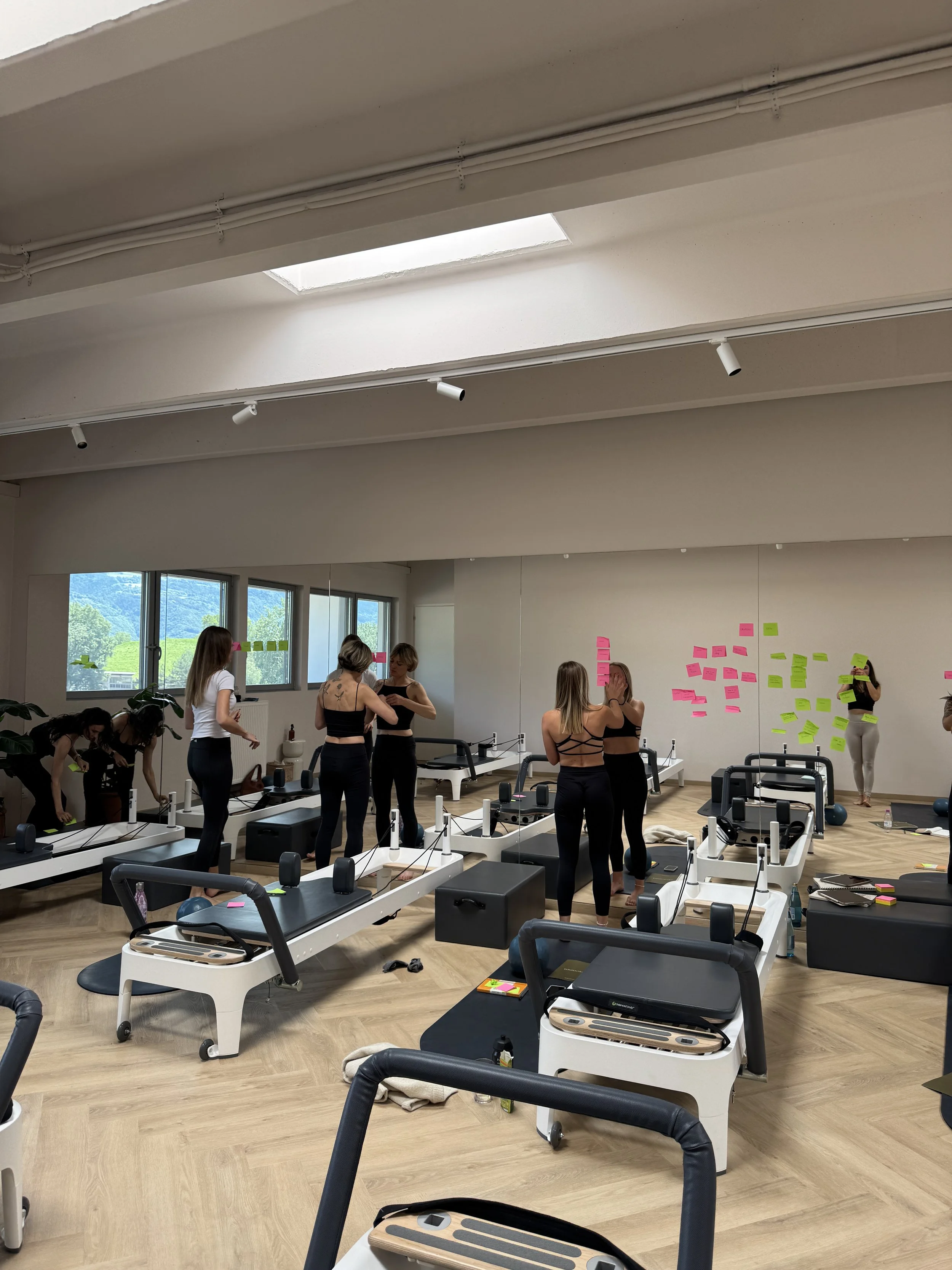 Ein Pilates-Studio mit mehreren Pilates-Geräten, Frauen in sportlicher Kleidung, die sich vorbereiten, und bunten Post-it-Notizen an der Wand.