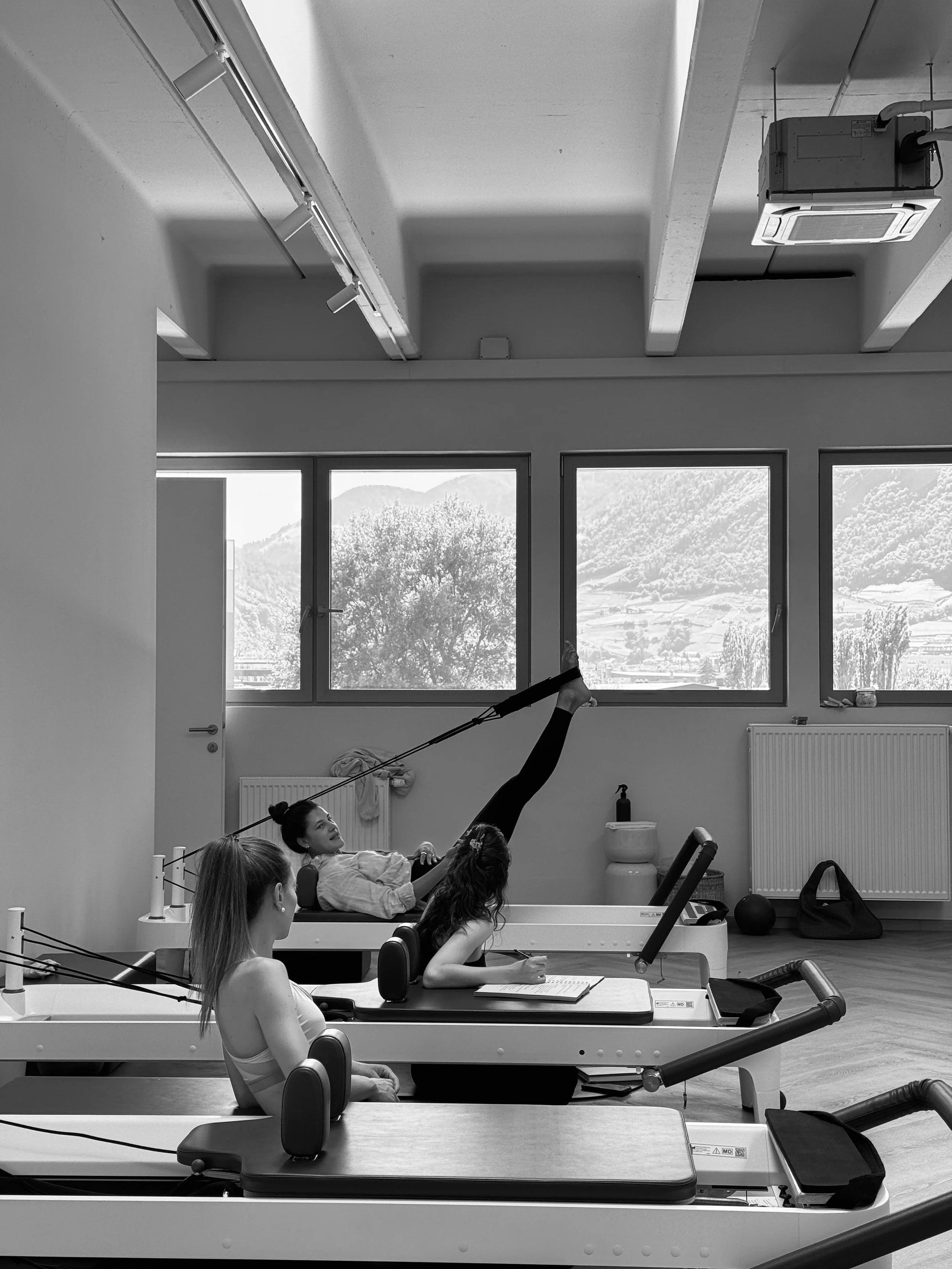 Drei Frauen trainieren im Pilates-Studio; eine liegt auf einem Reformpult, eine sitzt und eine liegt auf dem Rücken, während die Frau auf dem Reformpult eine Beinübung mit einem Widerstandsband macht.