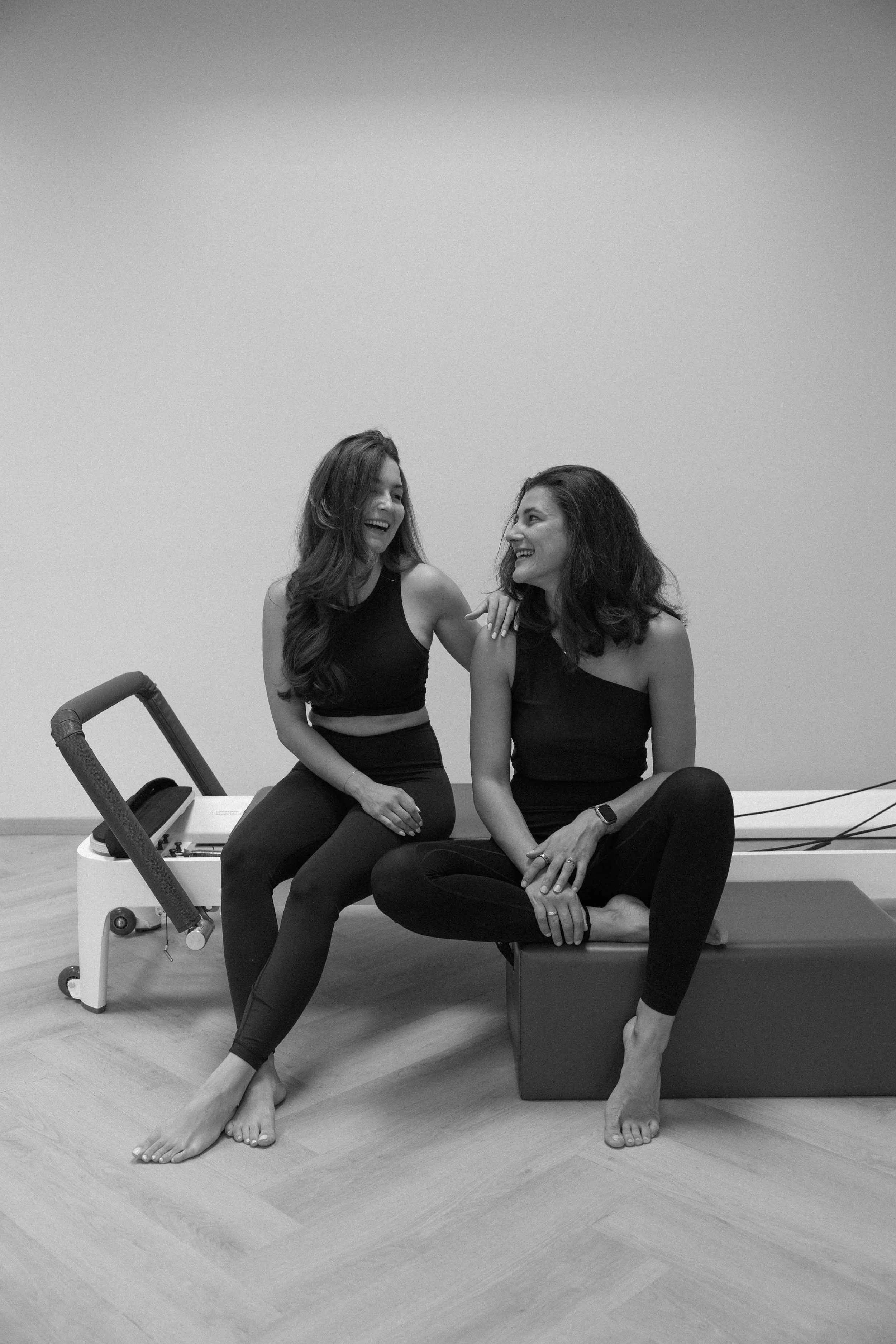 Zwei Frauen lachen in einem Raum mit Holzboden, eine sitzt auf einem Exercise-Box und die andere sitzt auf einer Pilates-Miege, beide tragen schwarze Sportkleidung.