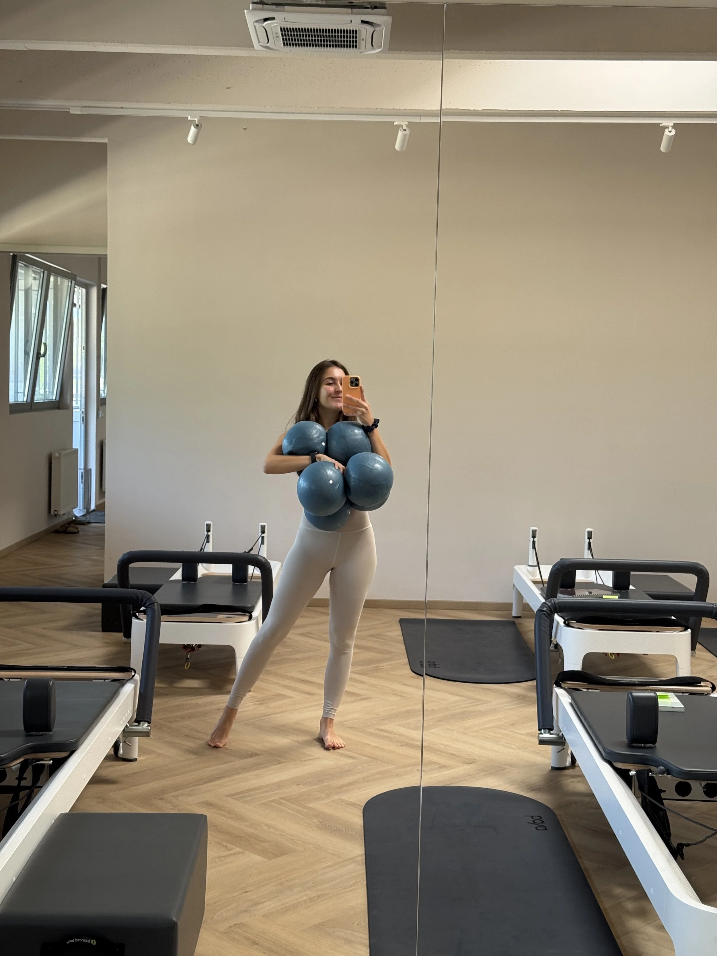 Eine junge Frau steht in einem Fitnessraum vor einem Spiegel und posiert für ein Selfie mit einem Smartphone. Sie trägt enganliegende beige Leggings und hält mehrere blaue Medizinbälle vor ihrer Brust.