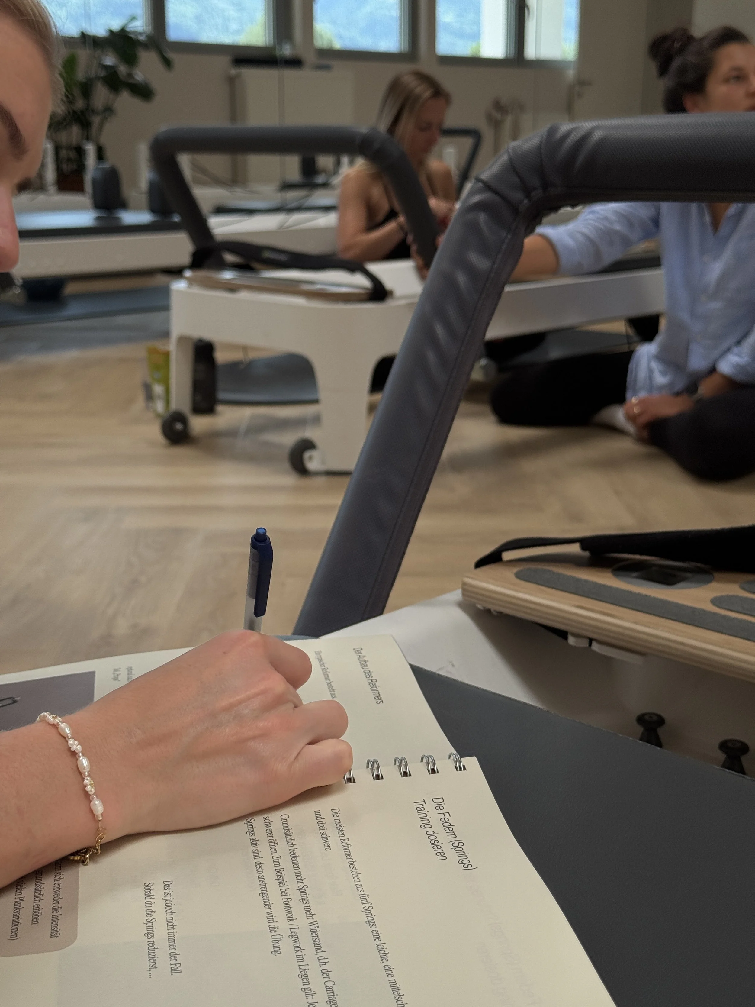 Menschen sitzen auf dem Boden in einem Raum mit großen Fenstern, während eine Person im Vordergrund eine Broschüre liest und schreibt.