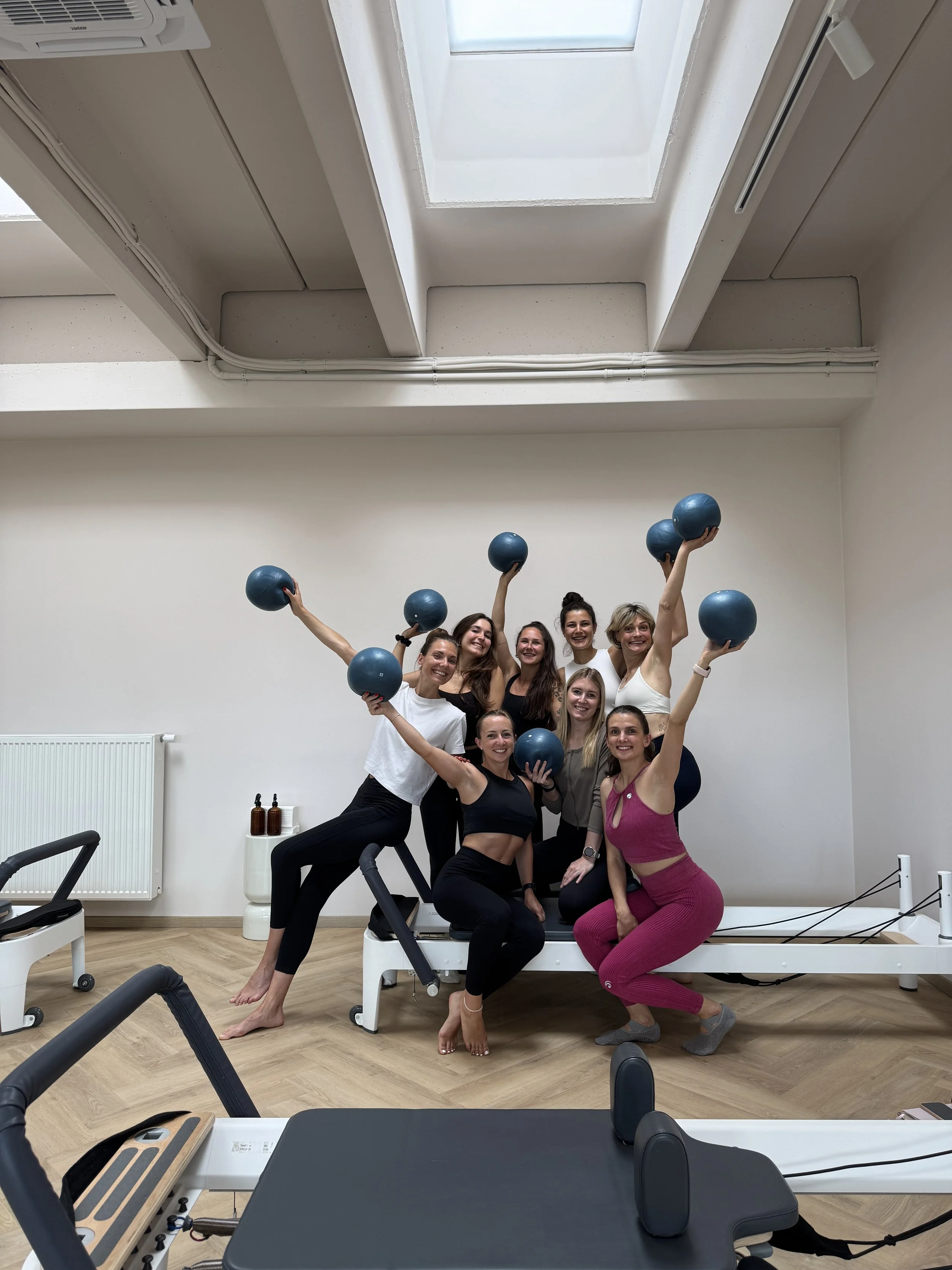 Gruppe von Frauen beim Pilates-Workout in einem Fitnessraum, einige halten blaue Medizinbälle, vor einem Pilates-Gerät, helle Wände, Holzboden, Dachfenster.