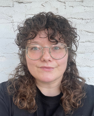 A photograph portrait of Ginger Carlson, wearing silver rimmed glasses with curly medium brown hair and a black shirt.