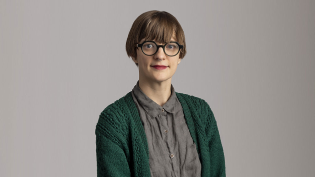 A photograph portrait of Brooke Leifso with short light brown hair, dark rimmed glasses, a nose piercing and a grey button top with a green sweater.