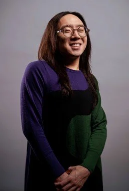 A photograph portrait of Sean Lee, wearing a colour-block top and glasses, with brown shoulder length hair.