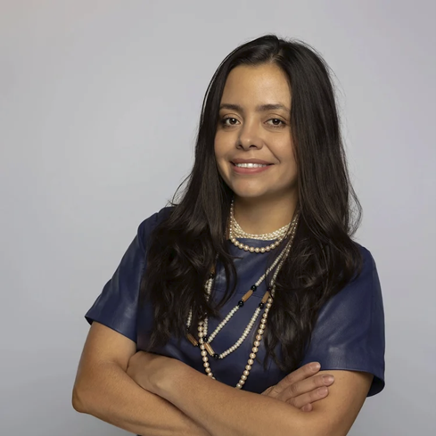 A photograph portrait of Nexi Alarcon, with long brown hair and brown eyes, wearing a blue top and several layered pearl and bead necklaces.