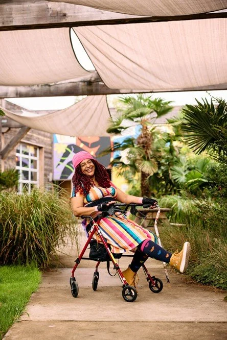 A portrait photograph of Claudia Alick sitting in a walker device wearing a multicoloured striped tunic top, a pink scarf, red braids, and colourful socks with heart shapes.