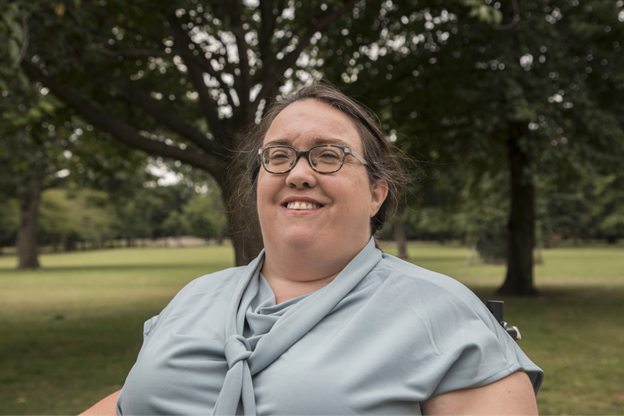 A photograph portrait of Louise Hickman, wearing glasses with brown hair and a silver grey blouse top with a tie at the front.