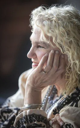 A photograph portrait of Carla Rice, with her hand near her face, with curly blonde hair and a blue and cream coloured top.
