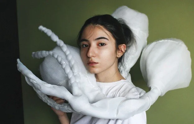 A photograph portrait of Aynaz Raoufian with dark brown hair and eyes, with white eyeliner, wearing a white top and holding a white textile sculpture wrapped around her shoulders.