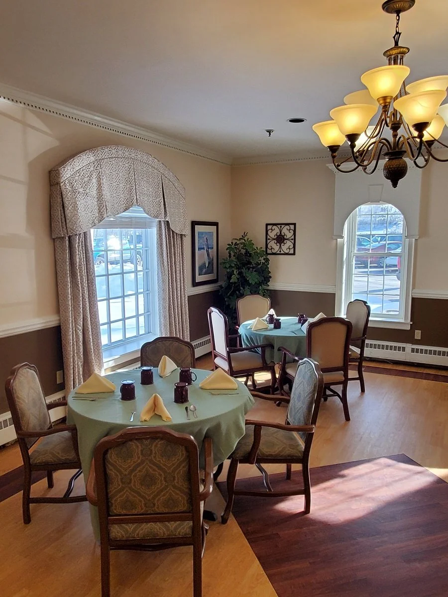 A dining room with two round tables set for a meal, each with six chairs, cream-colored walls, large windows, a chandelier, and a potted plant.