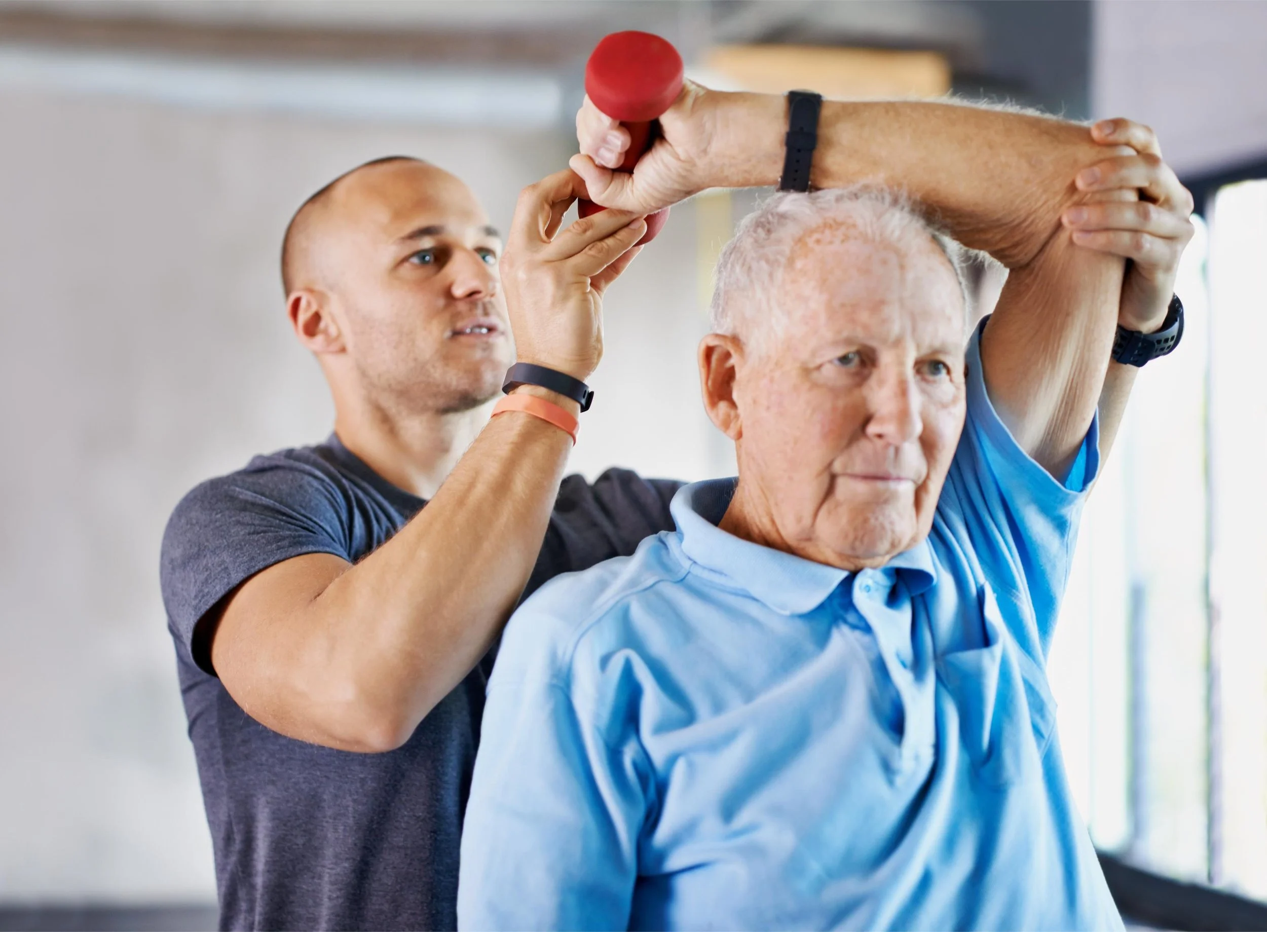 Younger man assisting elderly man with a light blue shirt during an exercise session, holding a red dumbbell above the senior's head in a gym or therapy setting.