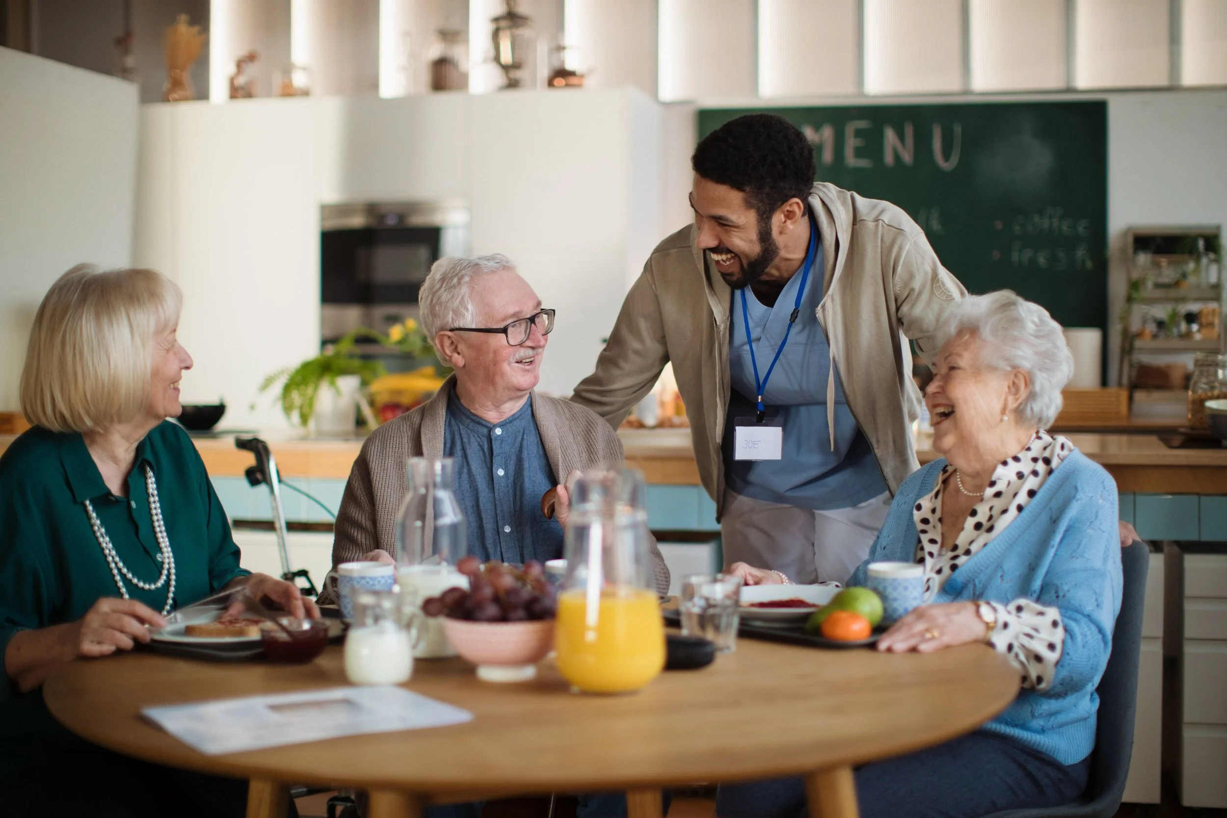 A young male caregiver with a name tag interacts with and serves breakfast to three elderly women at a table in a bright, modern kitchen. The women are smiling and laughing, enjoying a communal meal.