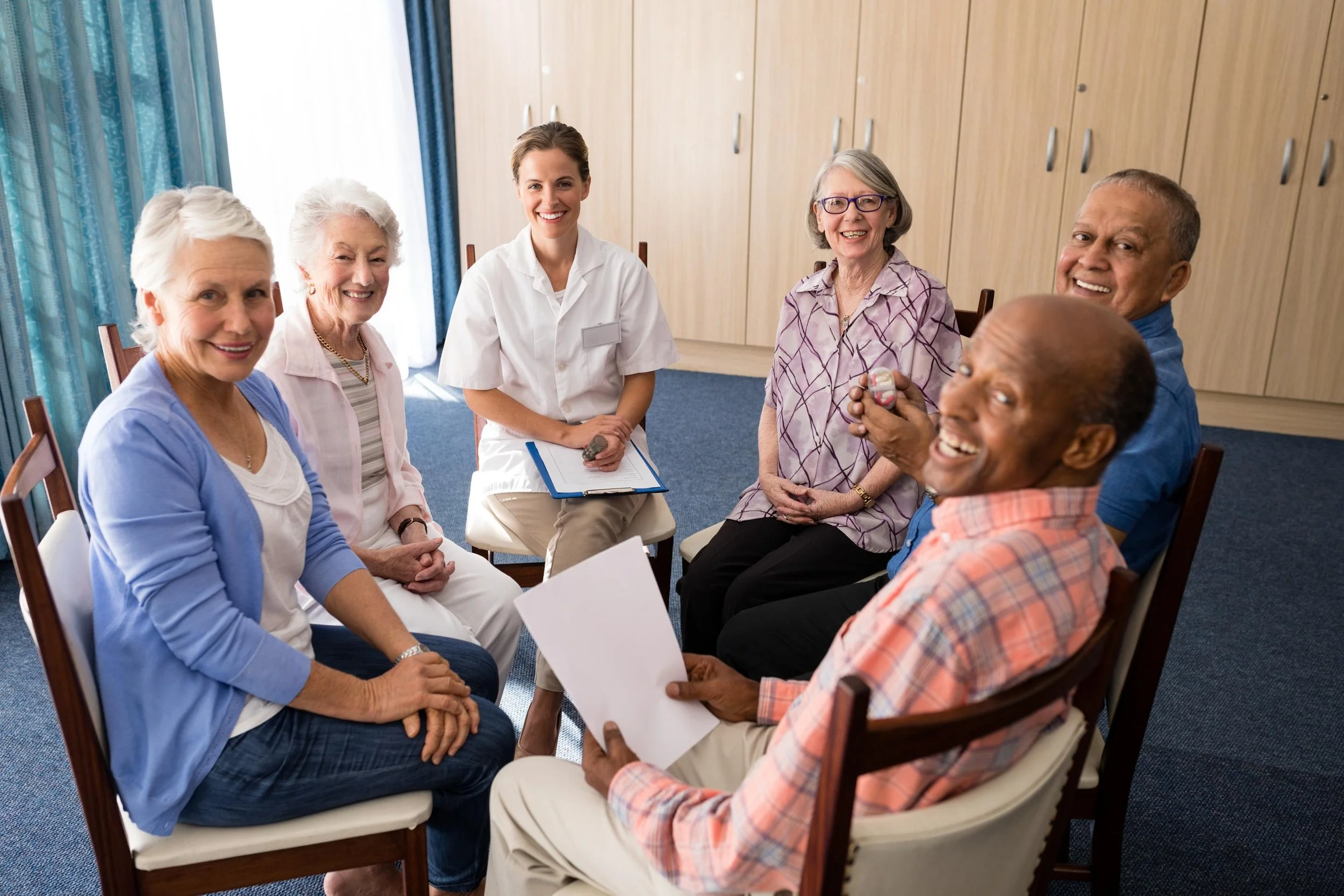A diverse group of six elderly and middle-aged people sitting in a circle in a room, smiling and engaging in a conversation, with a woman in healthcare attire holding a clipboard.