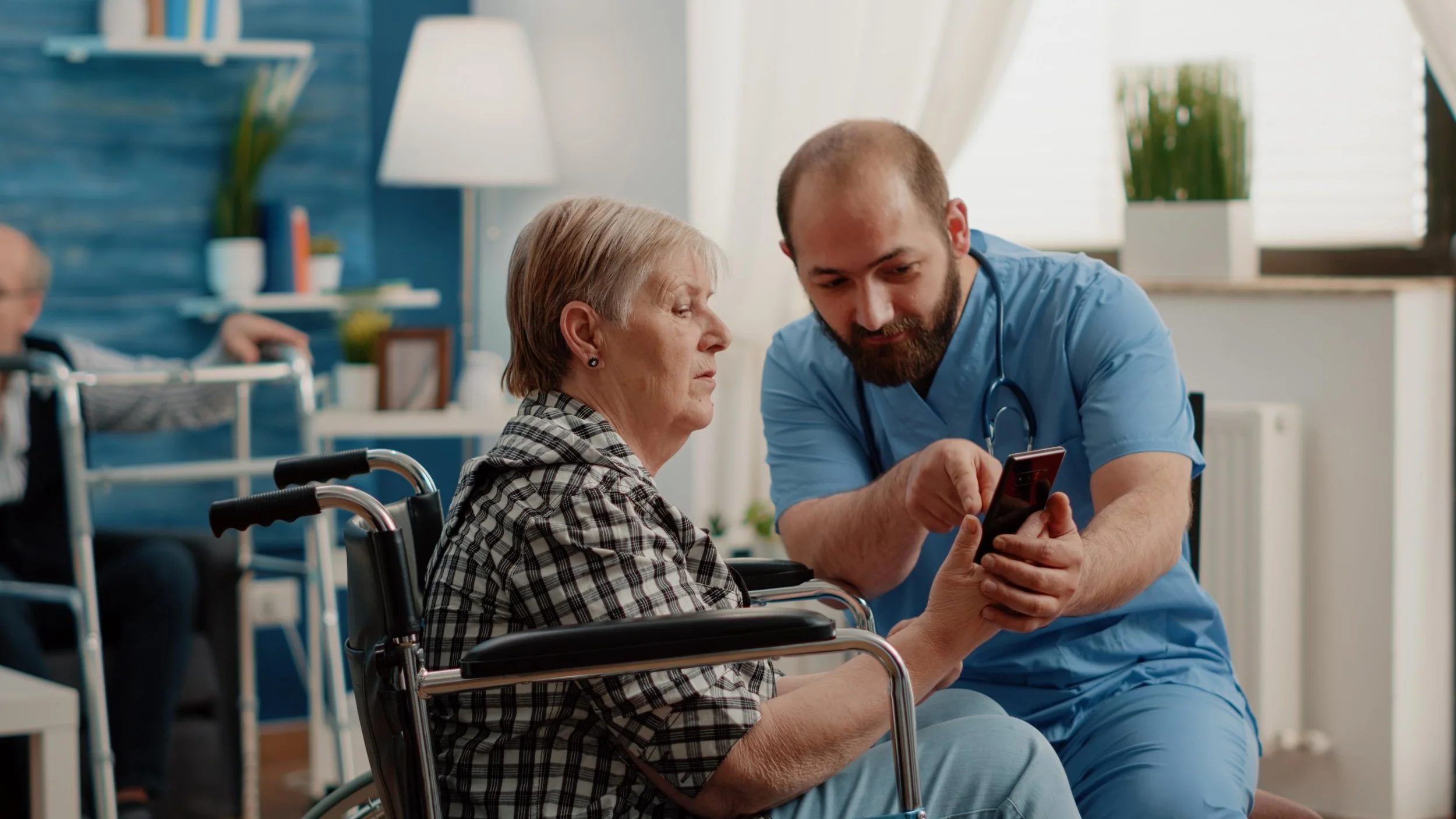 A healthcare professional in blue scrubs shows a smartphone screen to an elderly woman in a wheelchair in a bright, modern care facility with other patients in the background.