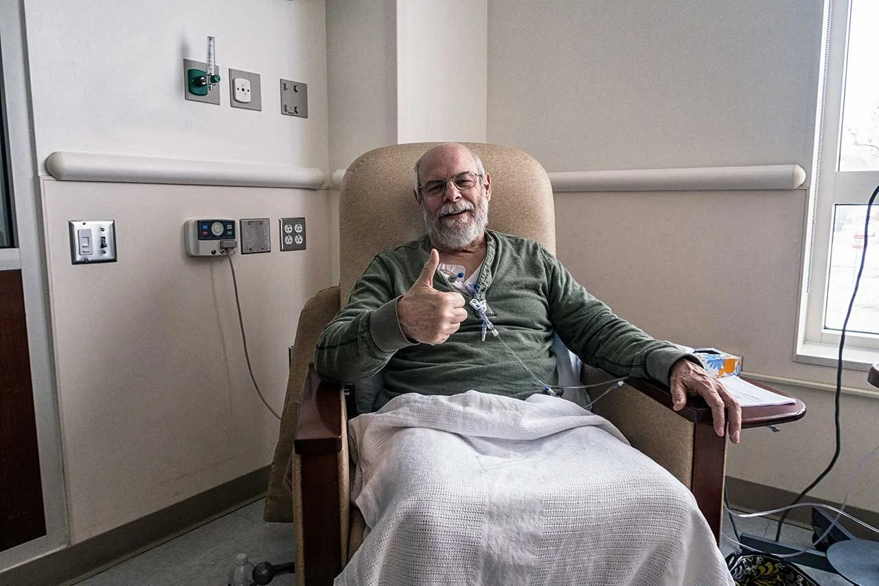An elderly man with glasses and a beard sitting in a hospital chair, giving a thumbs-up and smiling.