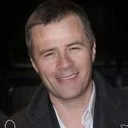 A smiling man with short hair, wearing a black shirt and blazer.