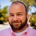 A man with a shaved head and beard, wearing a light pink shirt, smiling outdoors with trees in the background.