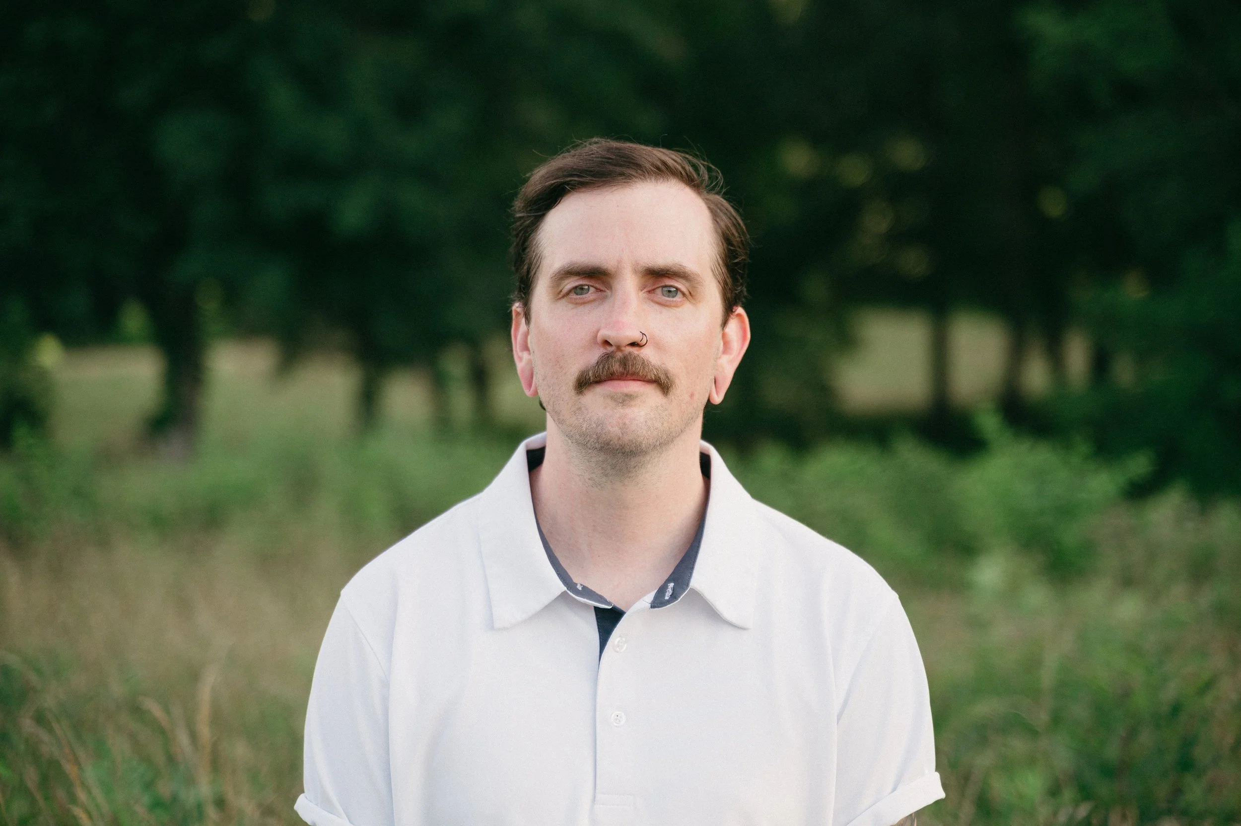 A man with a mustache and nose piercing standing outdoors in front of a blurred green forest background.