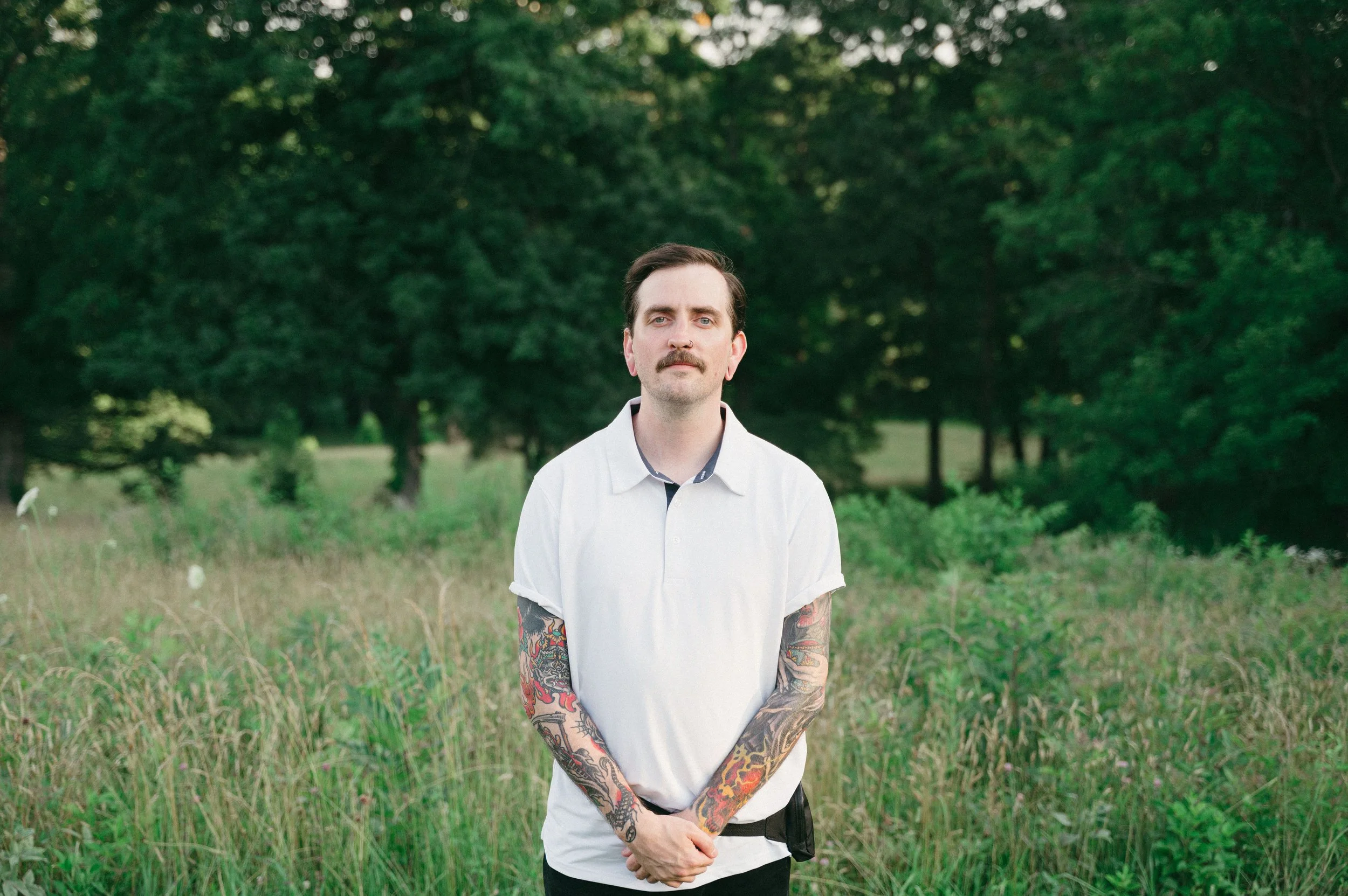 Young man with tattoos on arms, dark hair, and mustache standing in a grassy field with trees in the background.