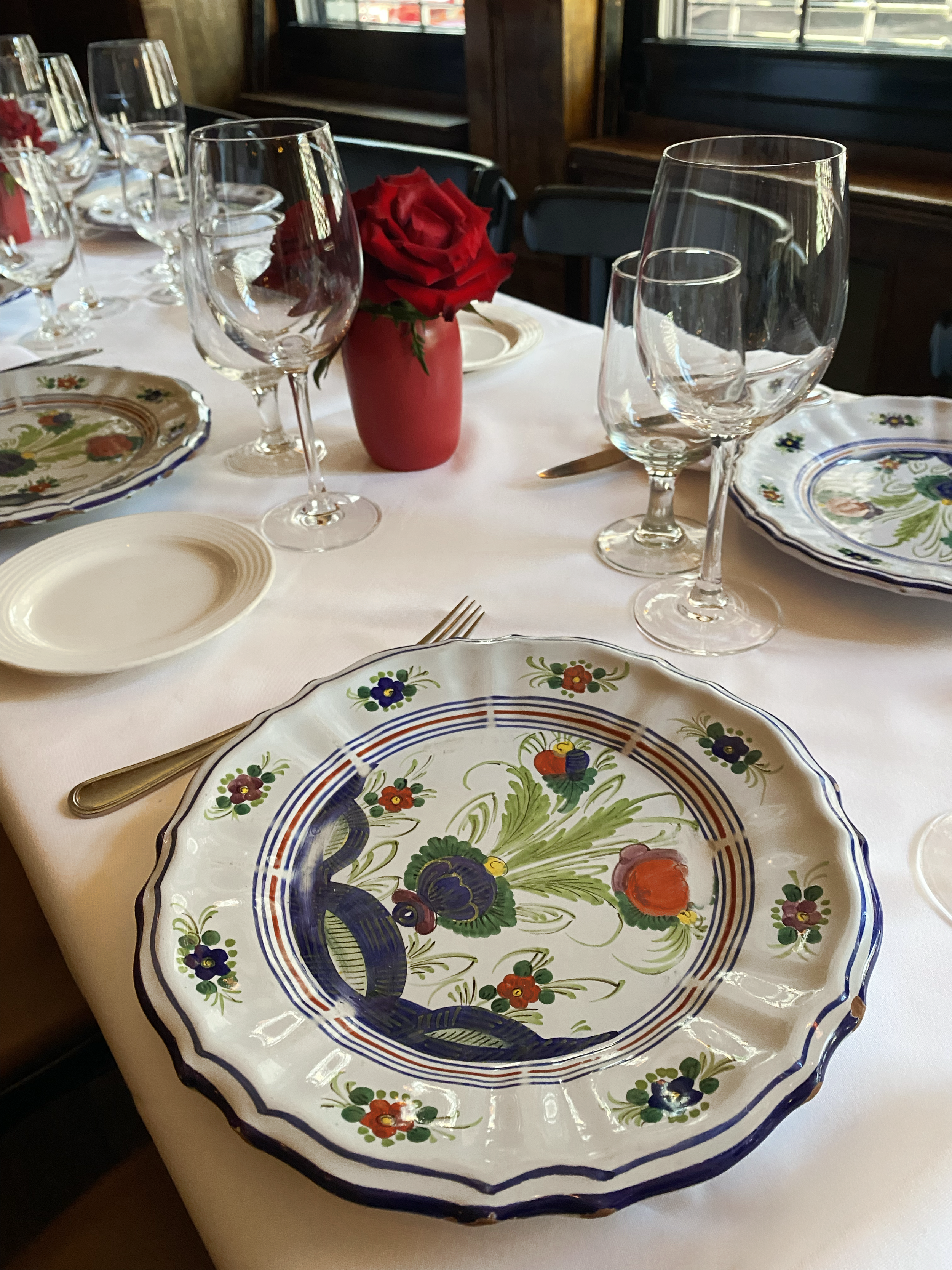 A dining table set with decorative floral plates, wine glasses, and a red rose in a red vase.