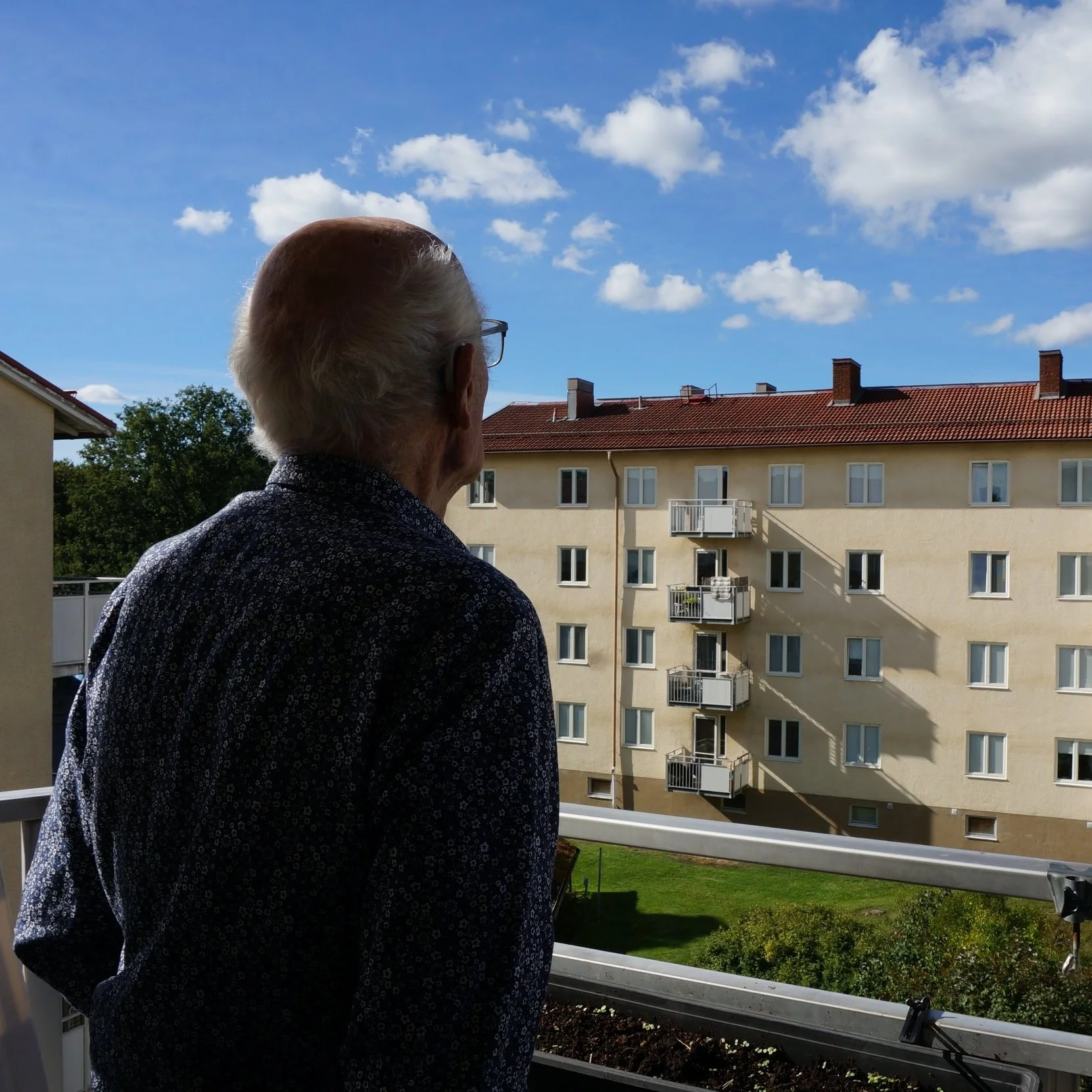 En äldre man med glasögon, som tittar ut från en balkong, mot en fyravåningshus med flera balkonger under en klarblå himmel med några moln.