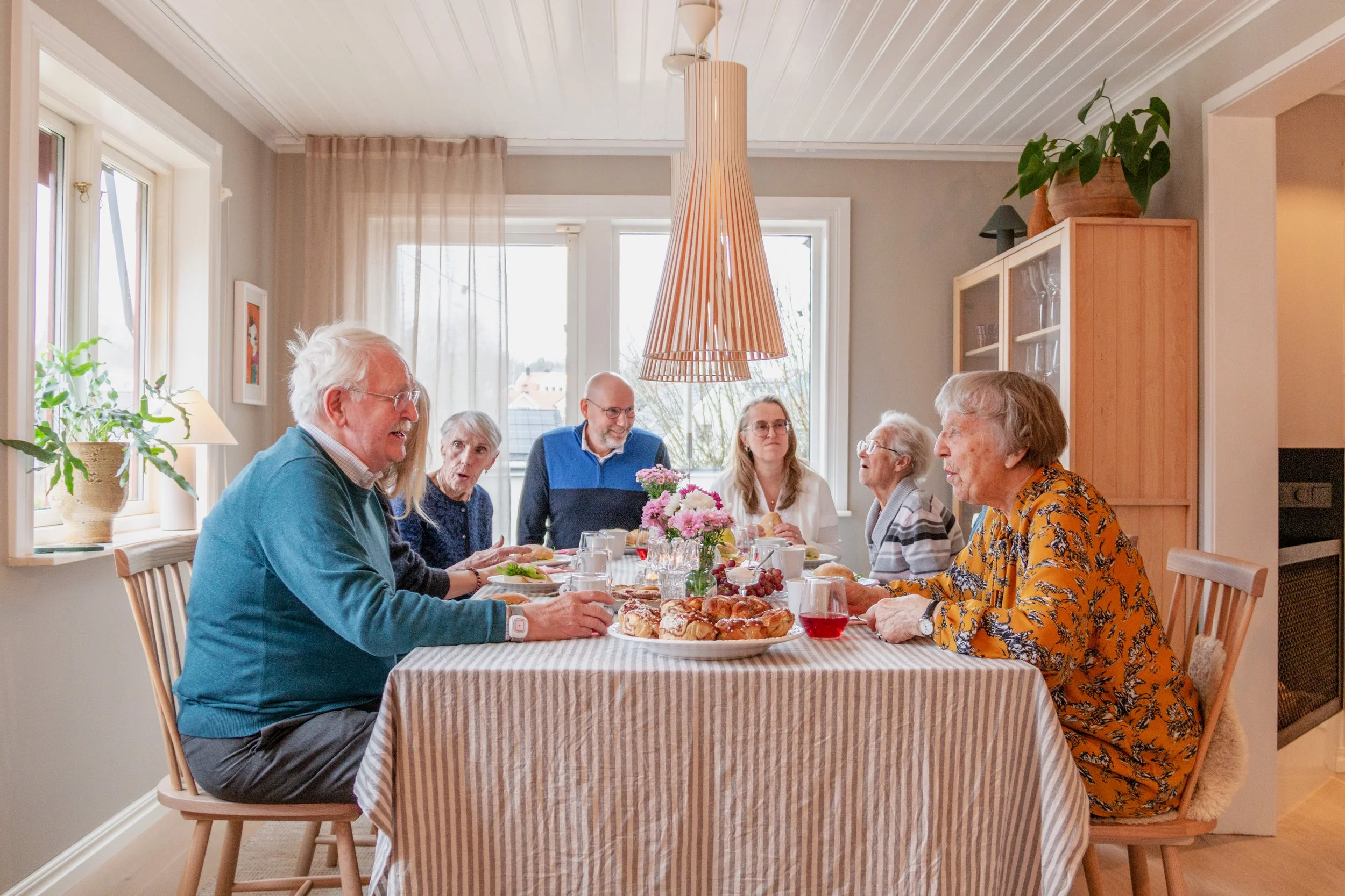Ett familjemåltid runt ett bord med mat och dryck i ett välbalkat rum. Sju personer av äldre och yngre åldrar samtalar och äter.