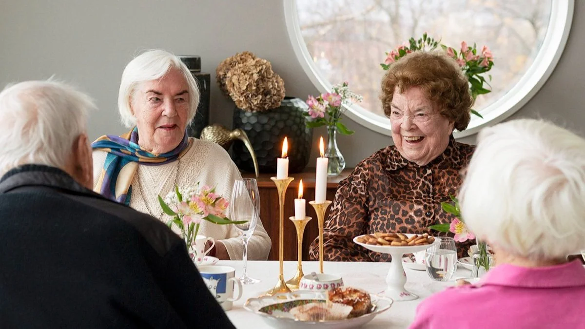 Ett bord med fyra äldre personer som sitter och pratar, dekorerat med blommor, ljus, kakor och te. Två kvinnor och två män, alla skrattar och har trevligt.