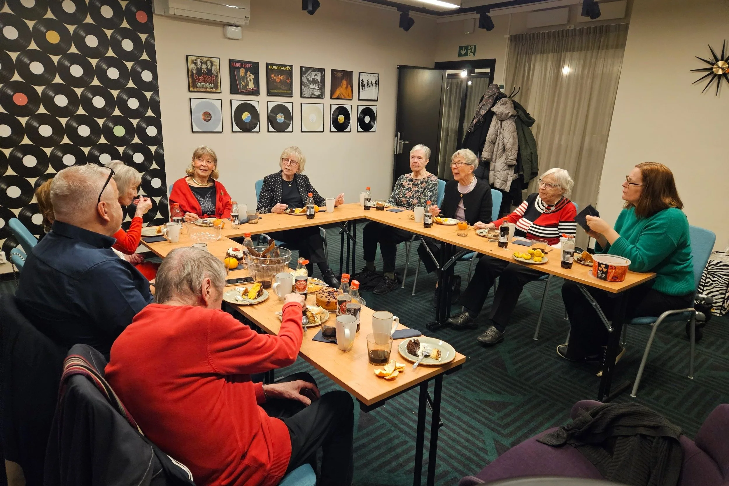 Ett gruppfoto av äldre personer som äter och samtalar runt ett bord under en sammankomst i ett sällskapsrum.