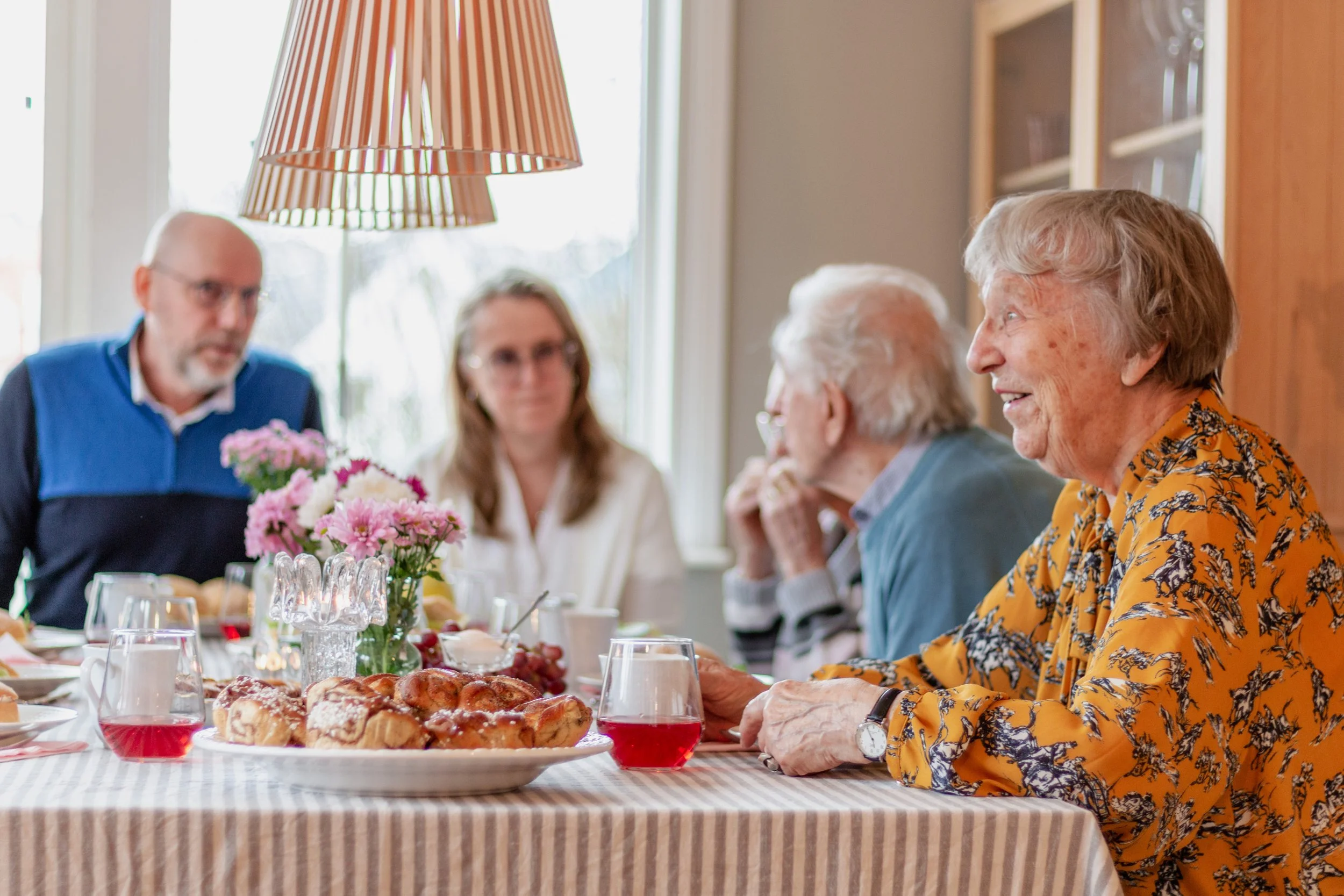 Eldre personer som äter och pratar vid ett bord med kaffe, kakor och blommor, i ett hem.