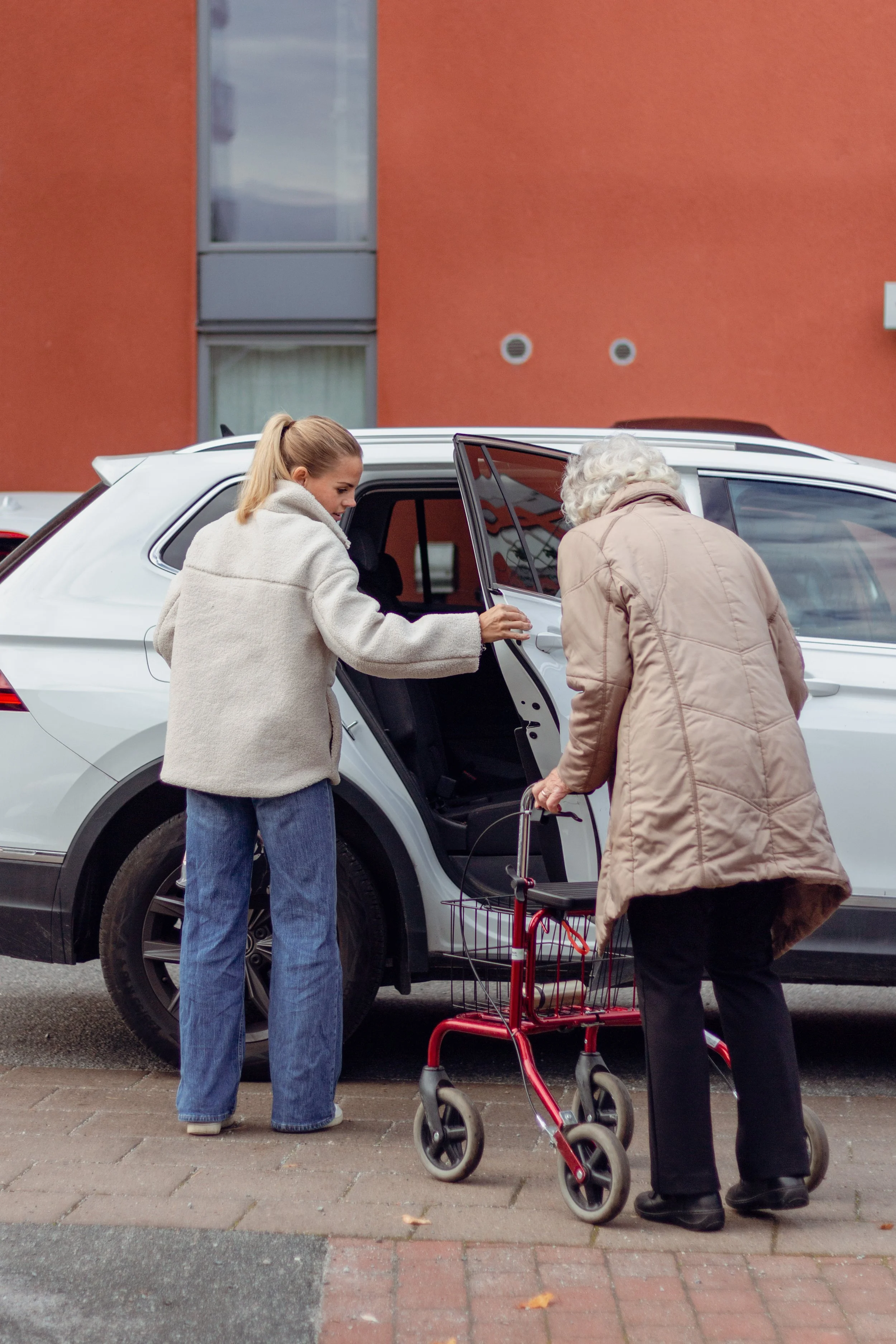Två kvinnor, en äldre och en yngre, som hjälper en person att komma i eller ur en vit bil med hjälp av en rollator, utanför ett bostadshus