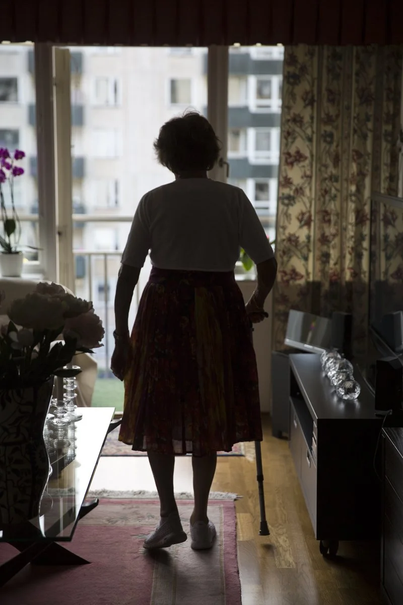 En person står inomhus framför ett fönster med utsikt över en stadsbyggnad, med ett gåstöd under armen.
