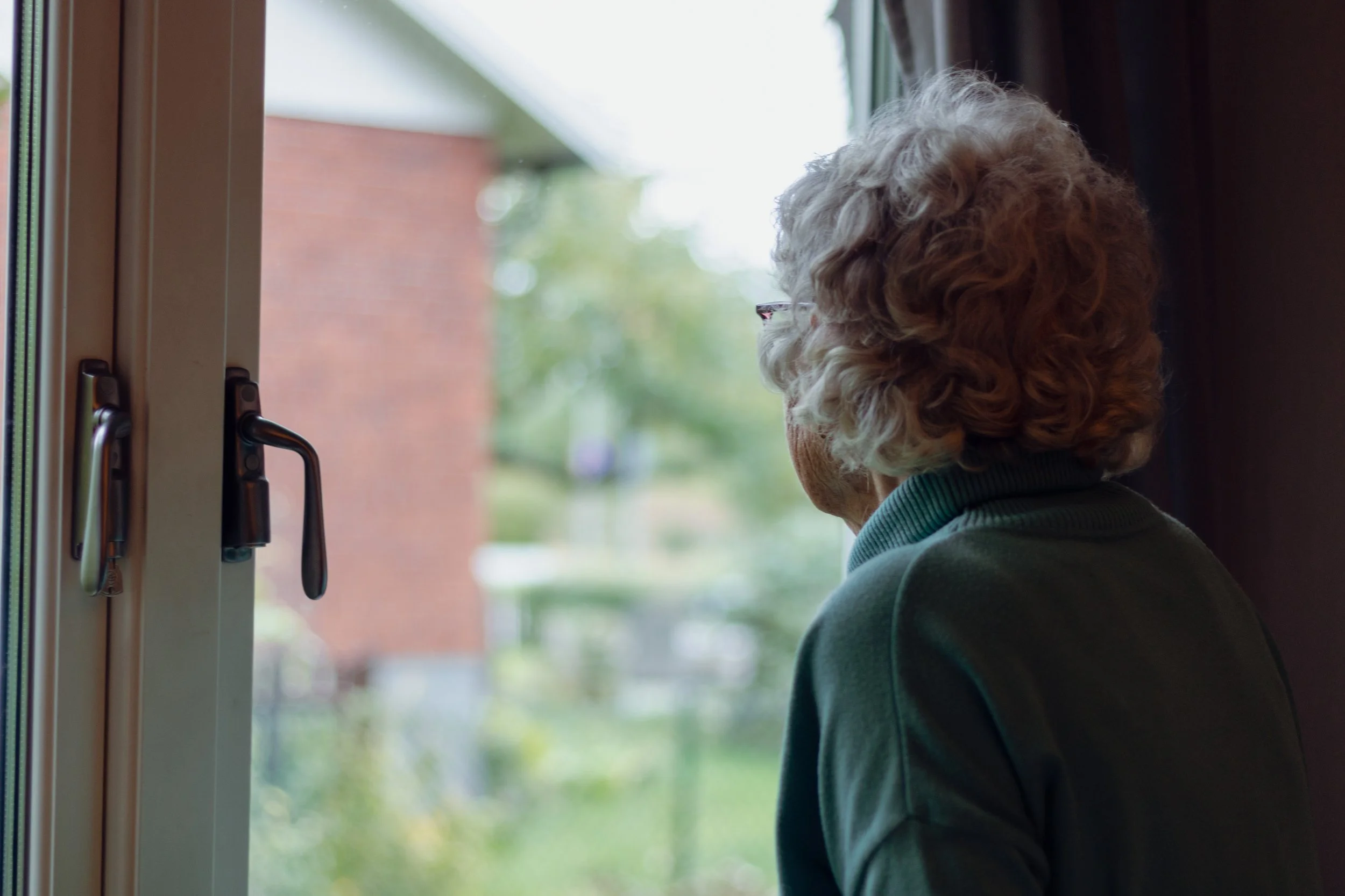 En äldre person med grå lockigt hår tittar ut genom ett fönster från insidan av ett hus eller en byggnad.