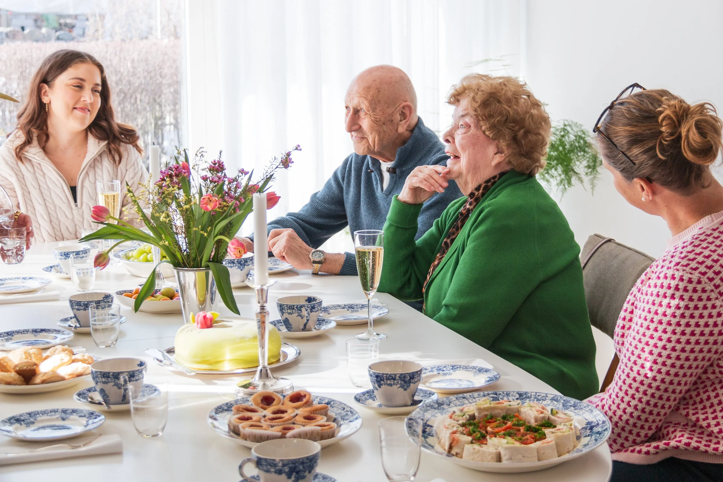 En grupp människor sitter vid ett matbord och äter tillsammans, de verkar ha en trevlig stund. Bordet är dekorerat med blommor och det finns olika sorters mat och drycker.