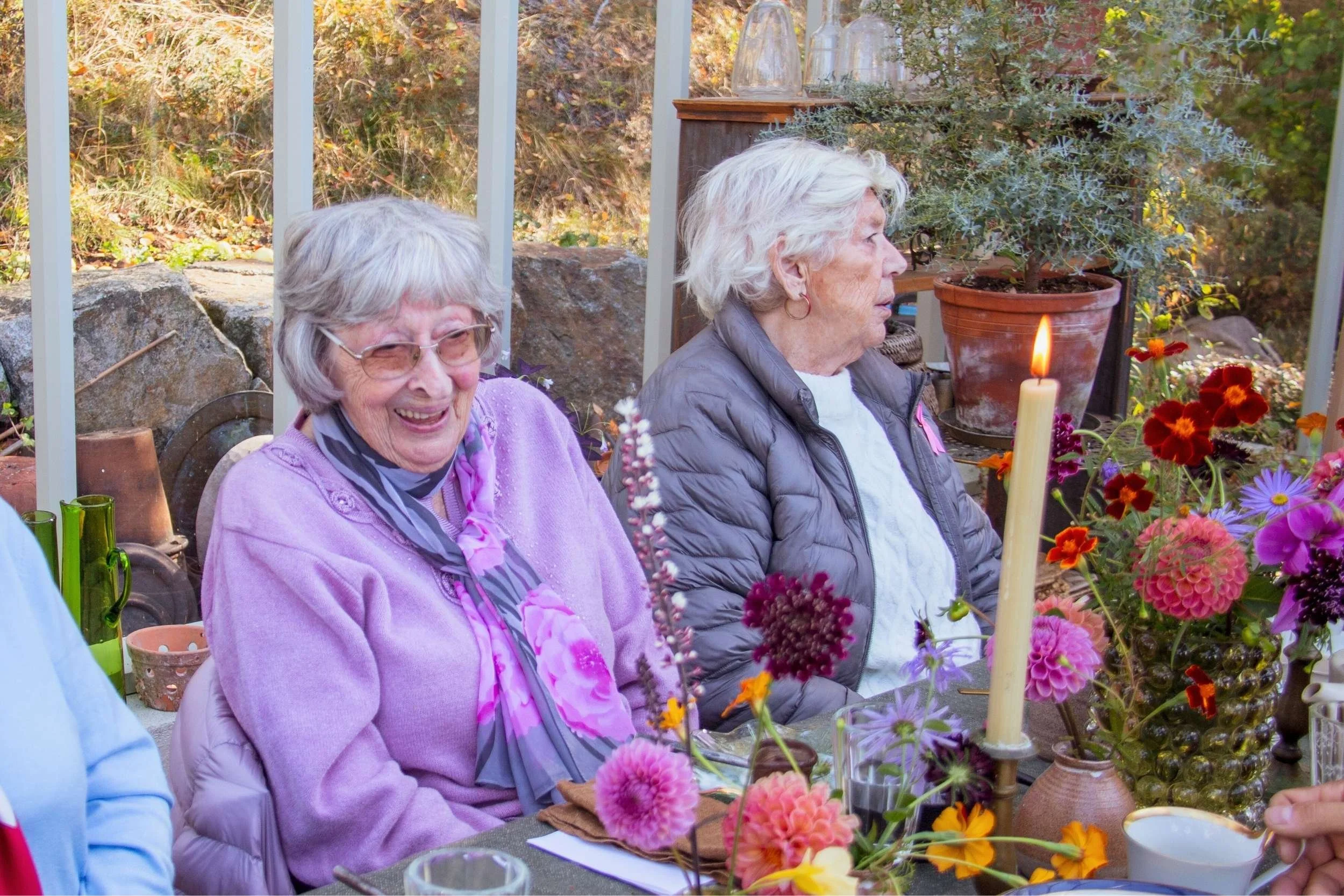 Två äldre kvinnor sitter vid ett bord inomhus, omgivna av parärer och blommor, en av dem är leende medan den andra sitter tyst.