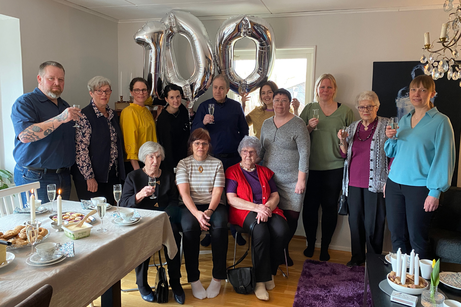 En grupp människor samlad för ett firande, några håller champagneglas, två stora folieballonger bildar siffrorna 100 i bakgrunden, på ett dukat bord finns mat och dryck.