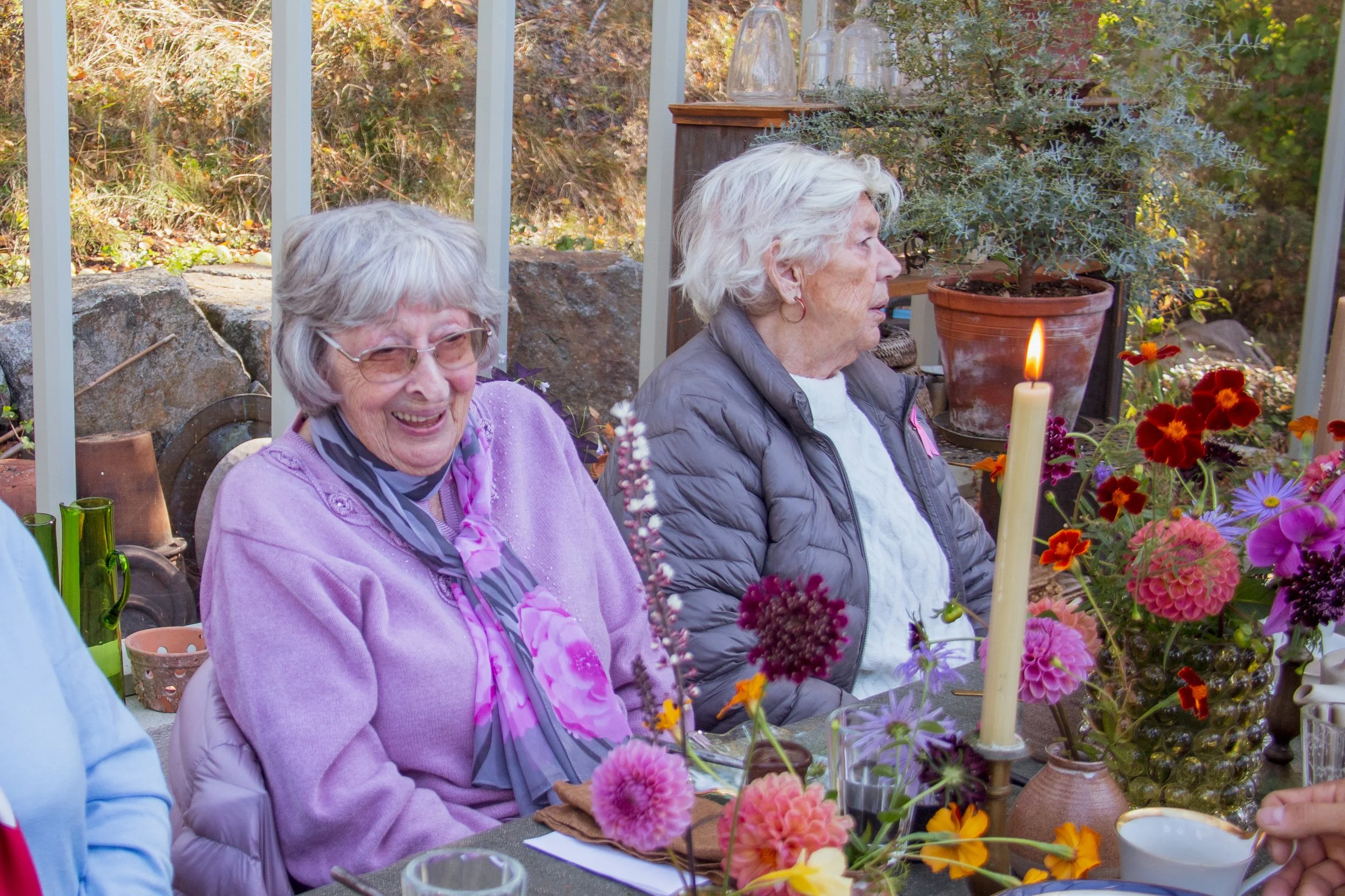 Två äldre kvinnor sitter vid ett bord med blommor, ett ljus och drycker i ett växthus eller uterum.