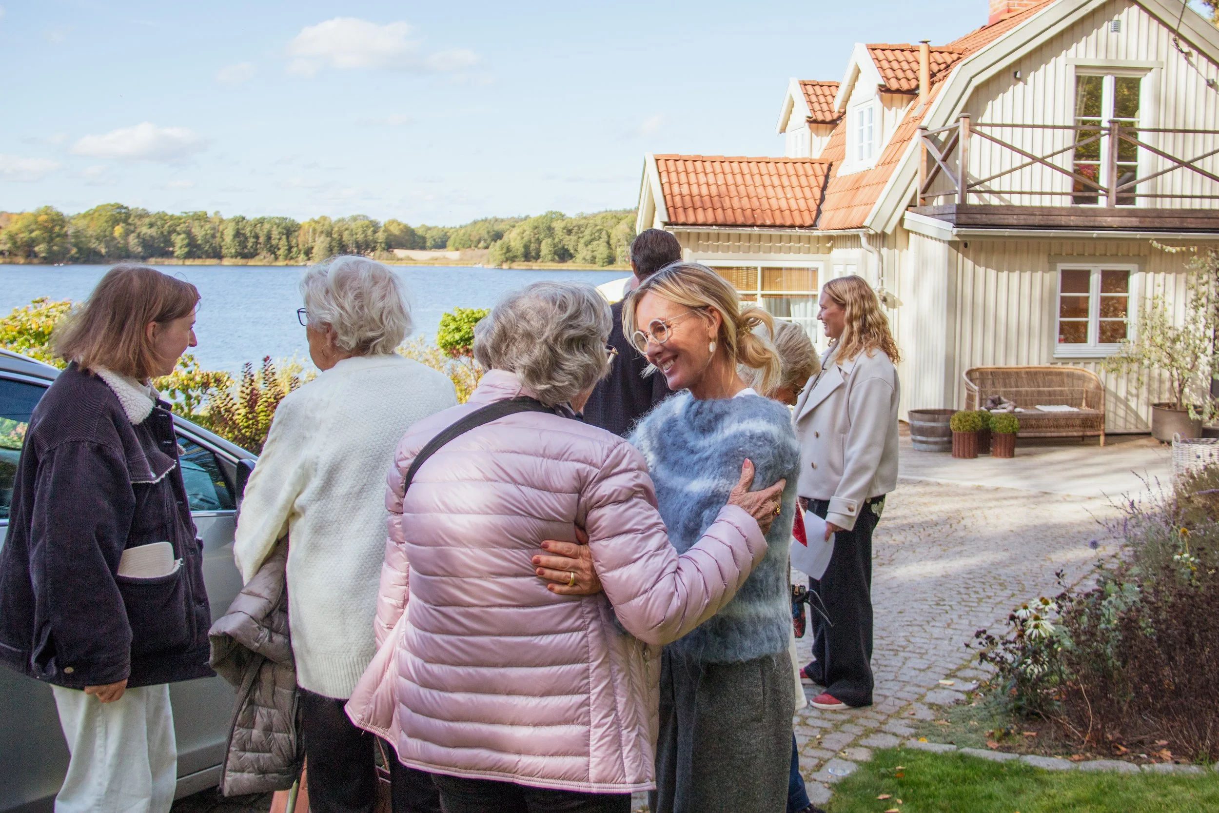 Flera personer som ler och pratar utomhus på en gräsmatta vid ett hus vid en sjö på höstdag