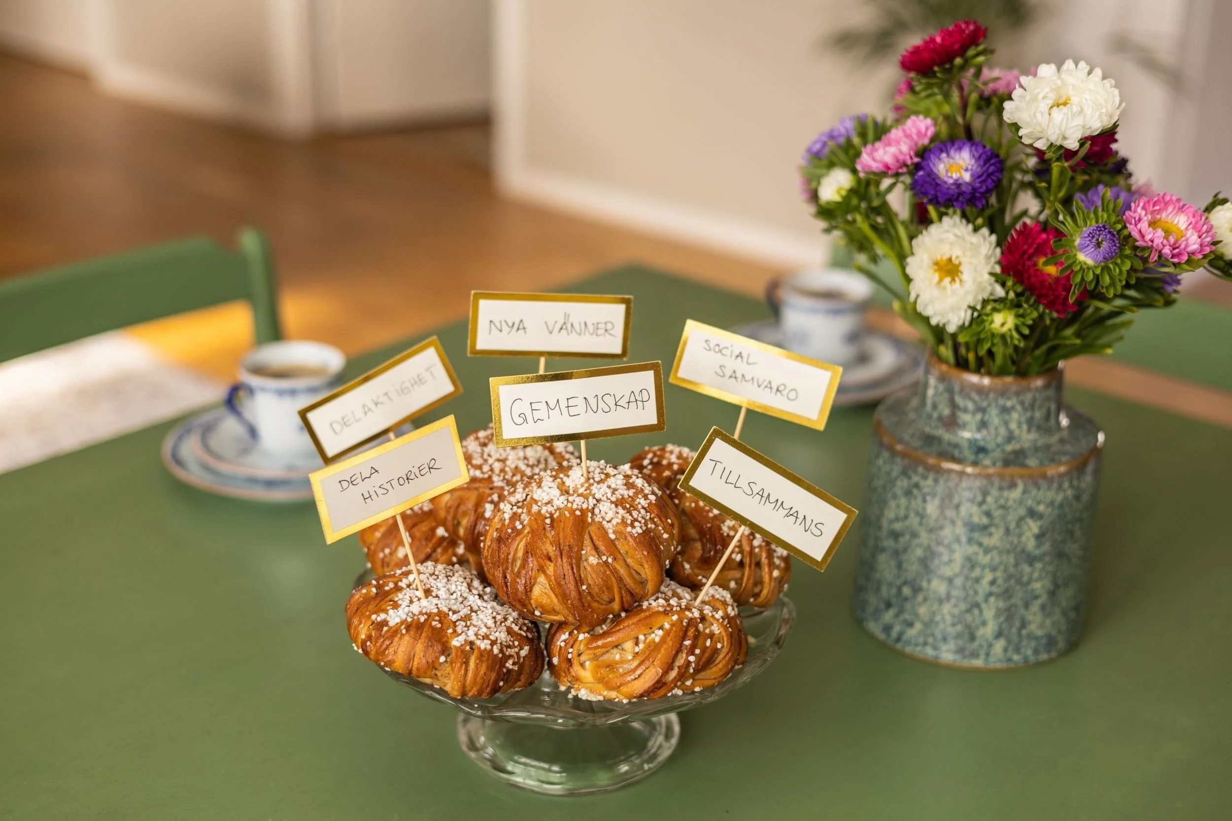 En skål med kanelbullar med olika små skyltar kring, ord som 'Gemenskap', 'Nya vänner', 'Social samvaro', 'Tillsammans', 'Delaktighet', 'Dela historier'.
