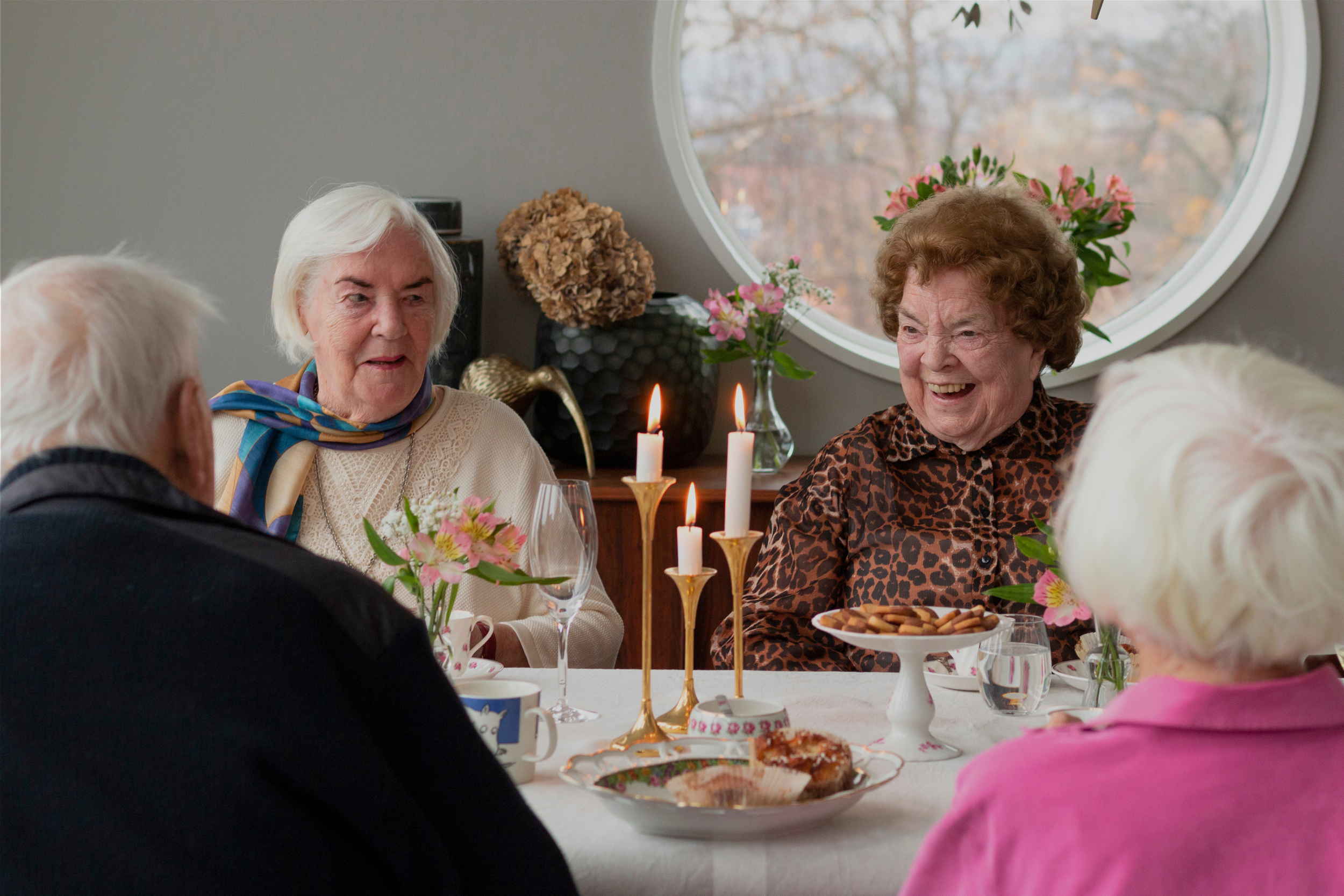 Äldre kvinnor och män som äter och pratar vid ett dukat bord med blommor, ljus och kakor i ett rum med ett fönster.