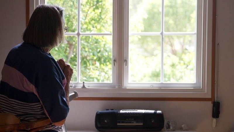 En äldre person sitter vid ett fönster och tittar ut, med en radio på fönsterbrädan.