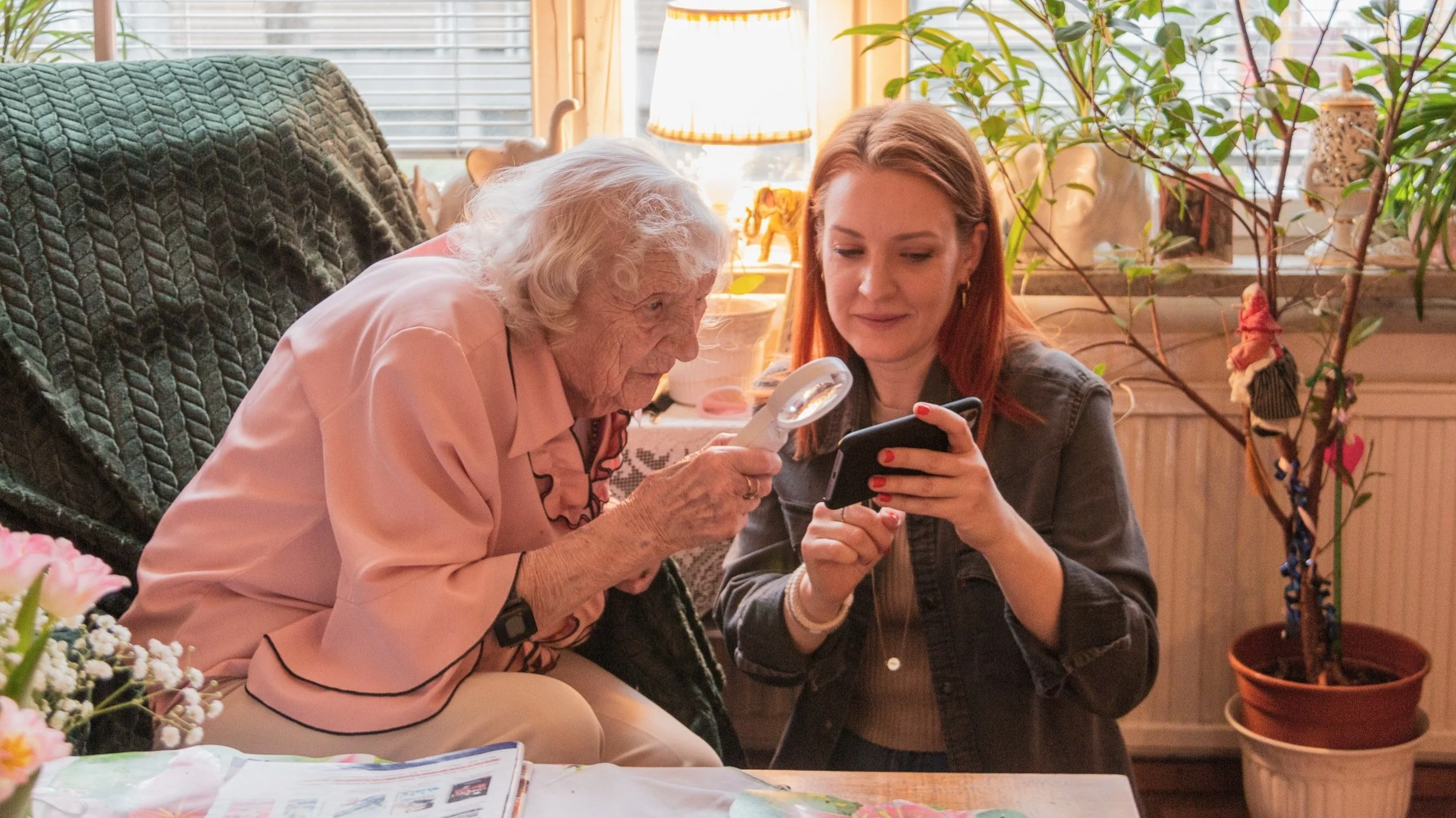 En äldre kvinna och en yngre kvinna tittar tillsammans på en smartphone i ett ljust rum med växter och dekorationer.