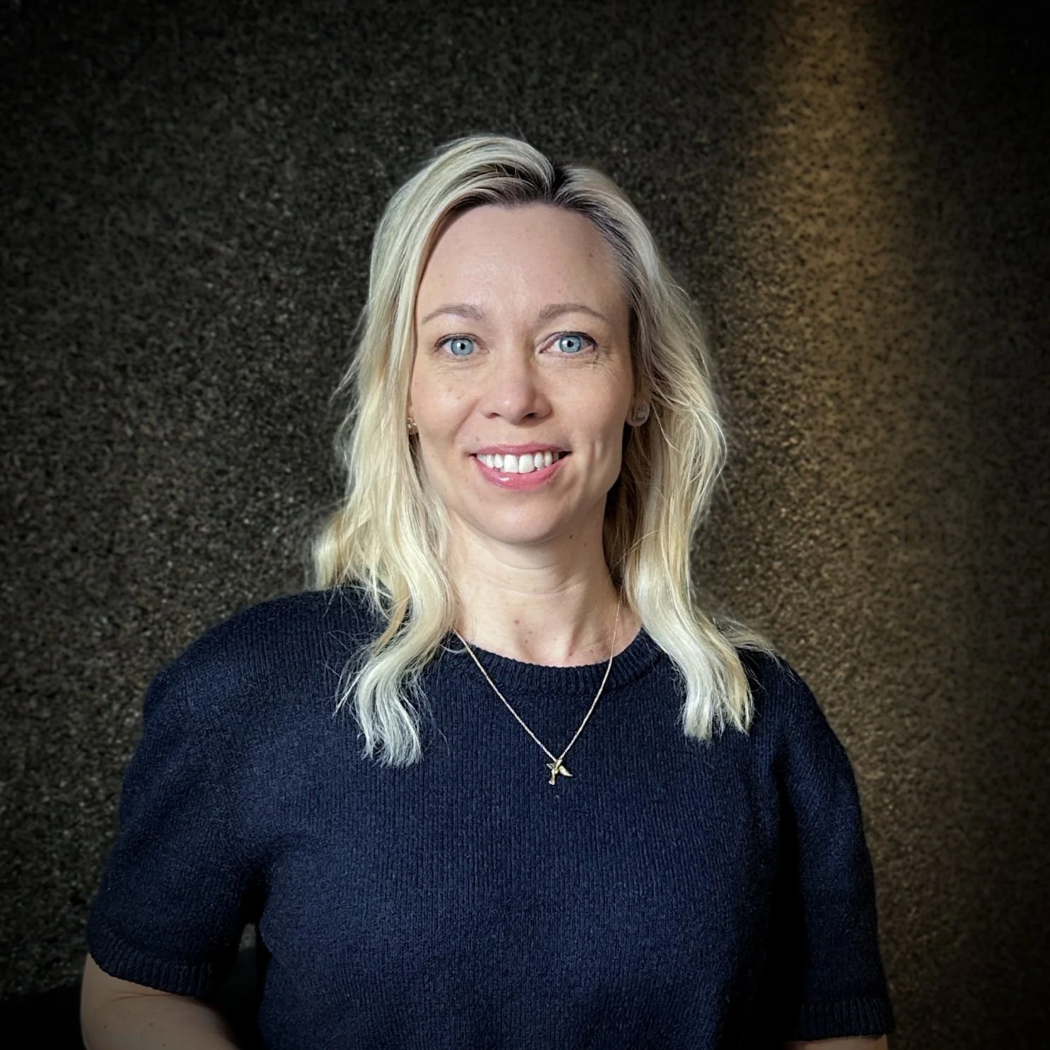 En kvinna med blont, lockigt hår, blå ögon och ett brett leende, bär en svart tröja och en guldhalskedja, står framför en mörk, texturerad bakgrund.