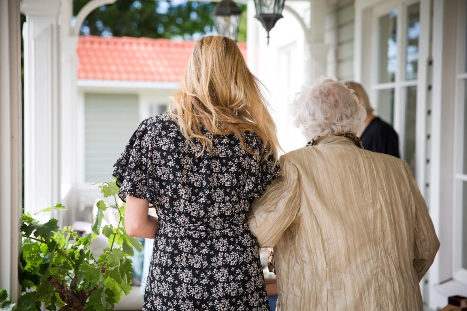 Två kvinnor, en yngre med rödbrunt hår och en äldre med vit hår, står nära varandra i ett fönsterparti och pratar, med ett vita hus med röd tak i bakgrunden.