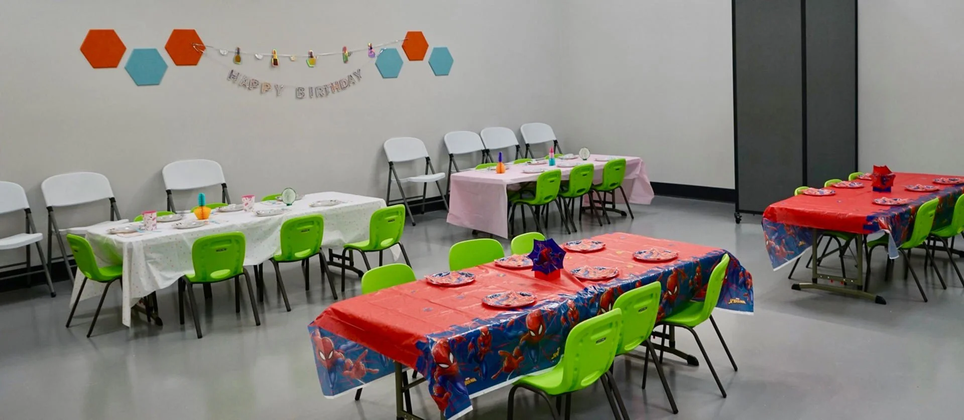 Room decorated for a child's birthday party with tables covered in colorful tablecloths, green chairs, and a birthday banner on the wall.