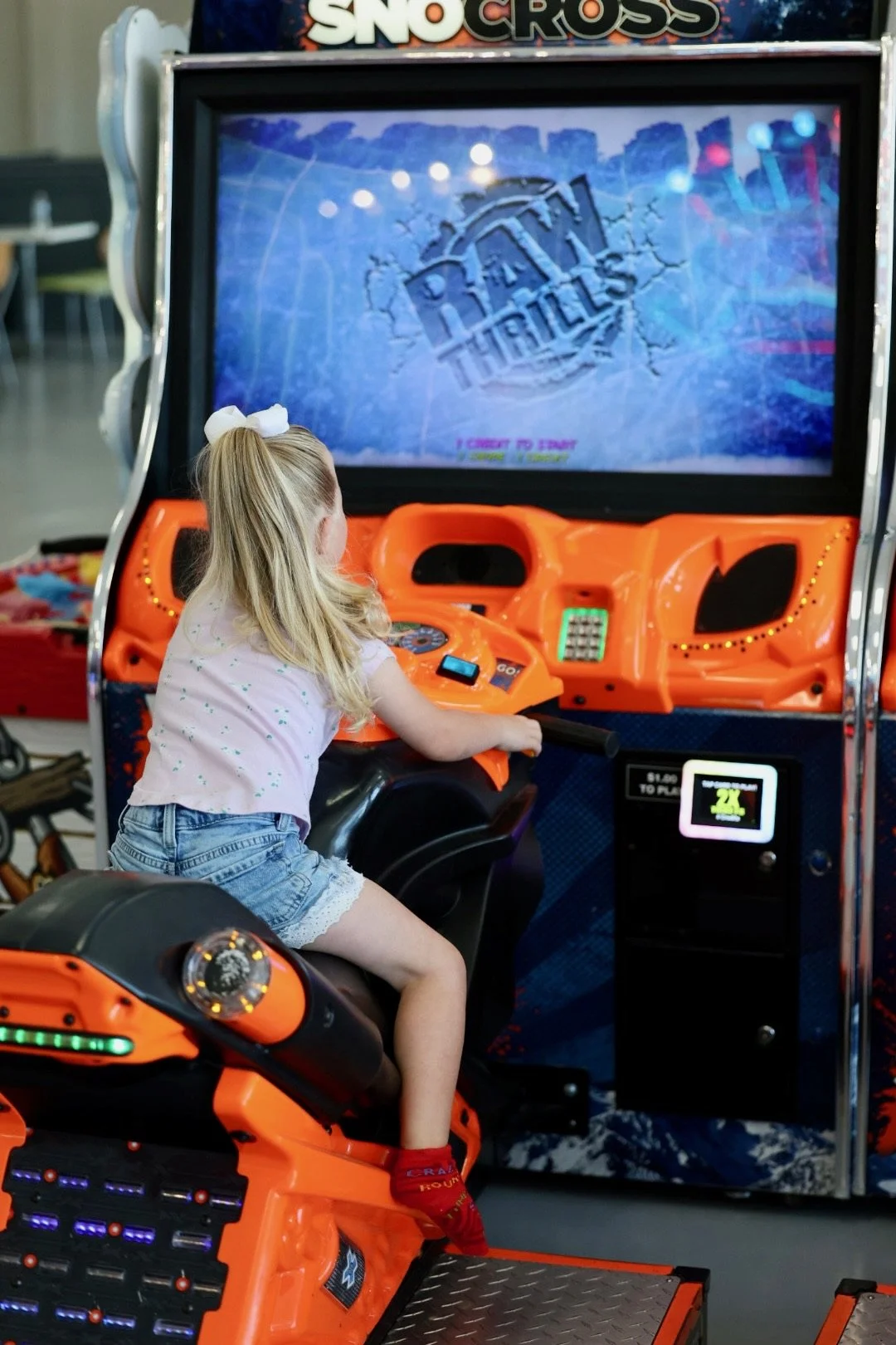 A young girl with blonde hair, wearing a white shirt, denim shorts, and red socks, is playing an arcade motorcycle racing game called 'SnoCross' at an arcade. She is sitting on an orange motorcycle seat controller, holding the handlebars, and facing 