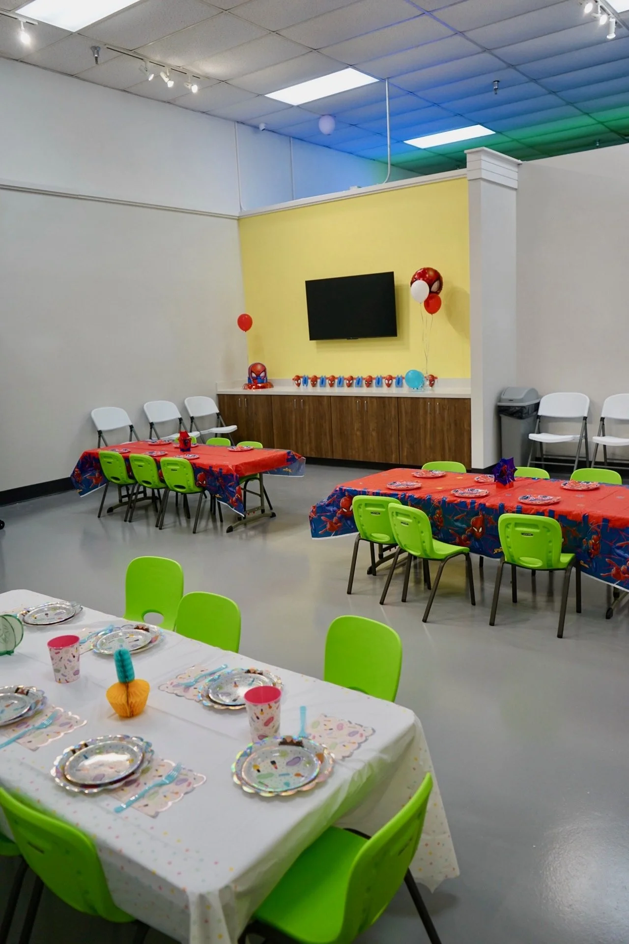 Party room decorated for a celebration with green chairs at a long table with a white tablecloth, colorful paper plates, cups, and a small pumpkin centerpiece. Decorations include balloons and Spiderman-themed items, with a yellow accent wall and a m